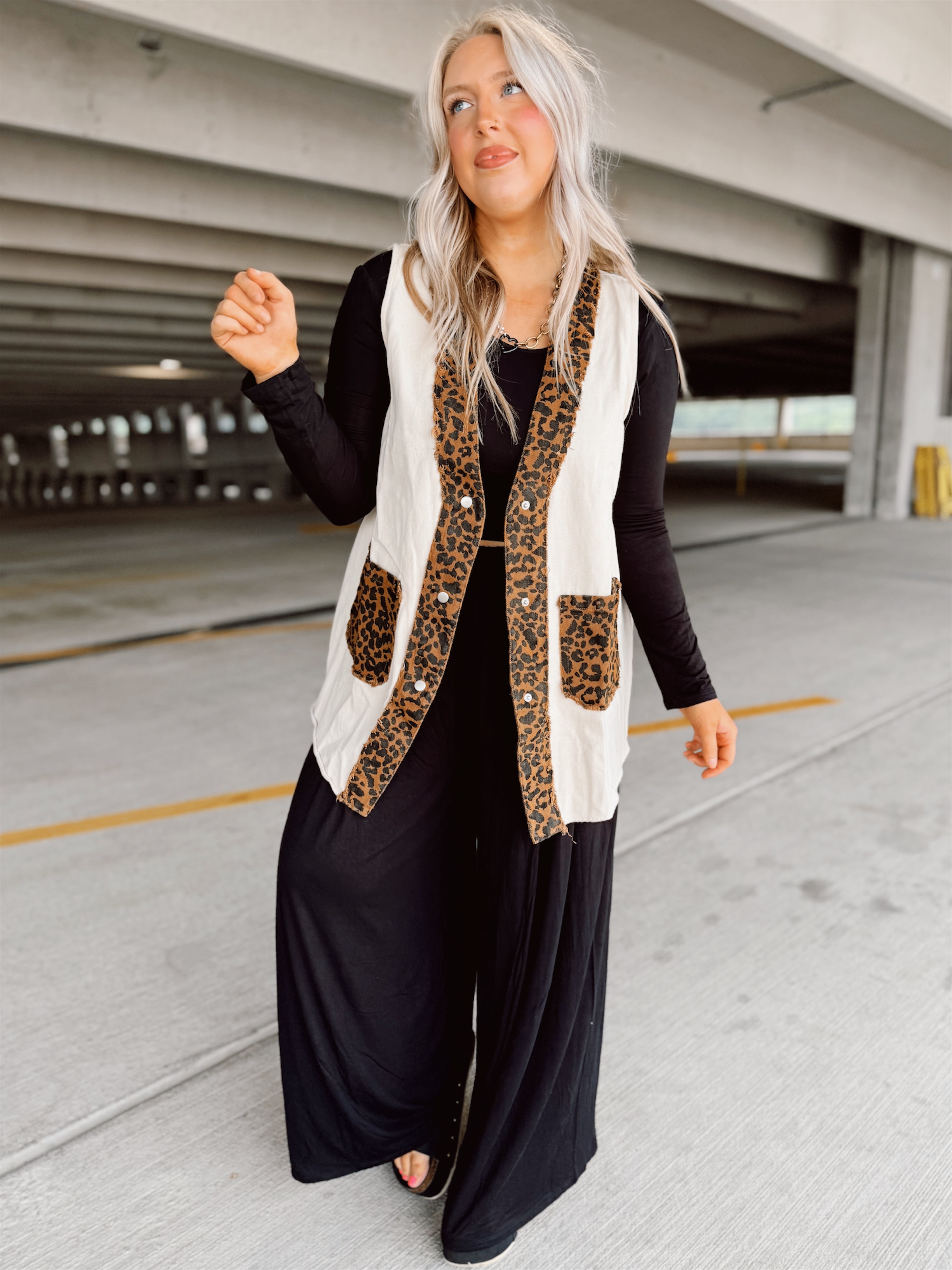 Woman wearing a leopard print vest over a black outfit in an indoor parking garage.