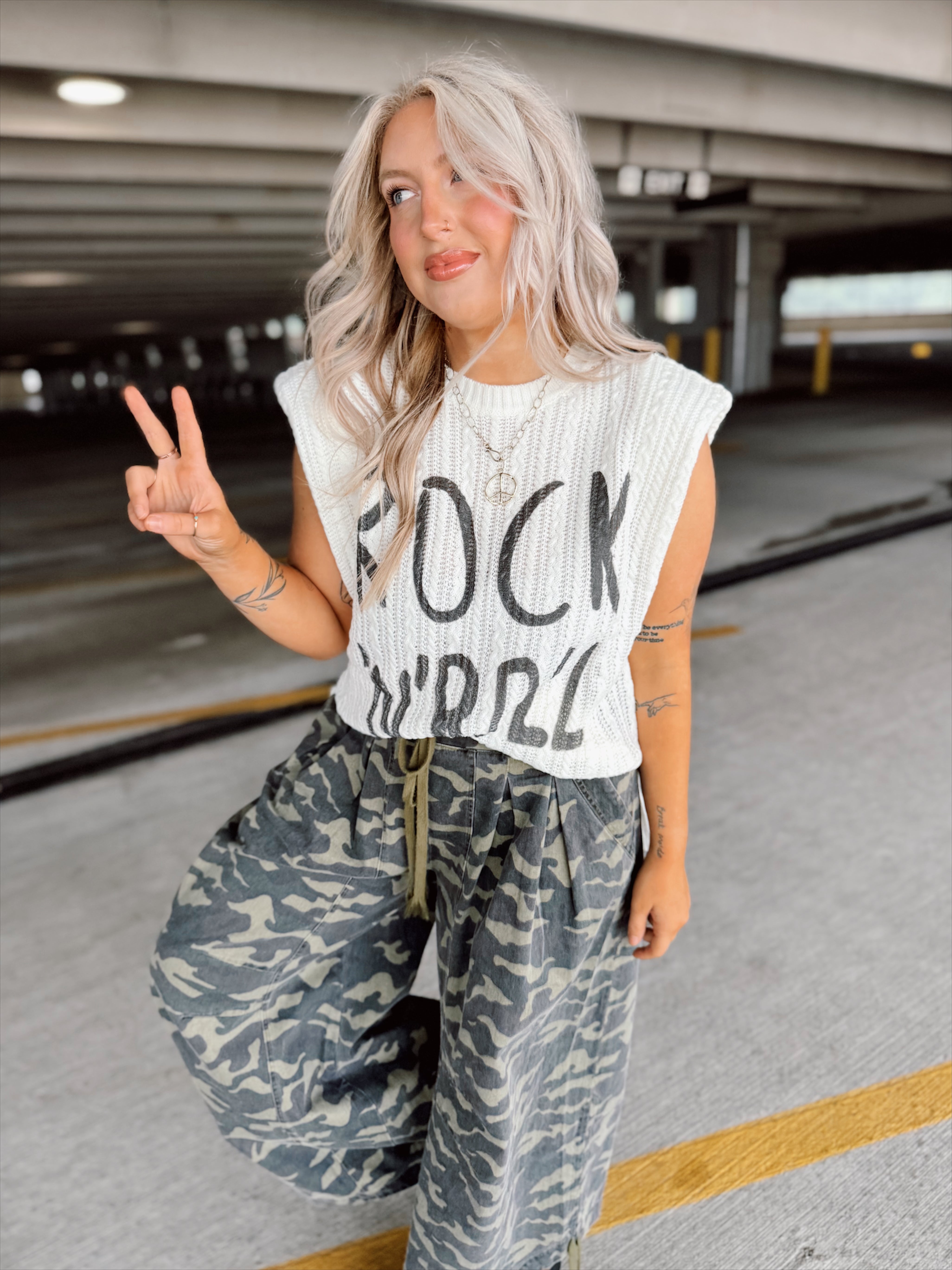 Woman in a parking garage wearing a sleeveless top with text and camouflage pants.