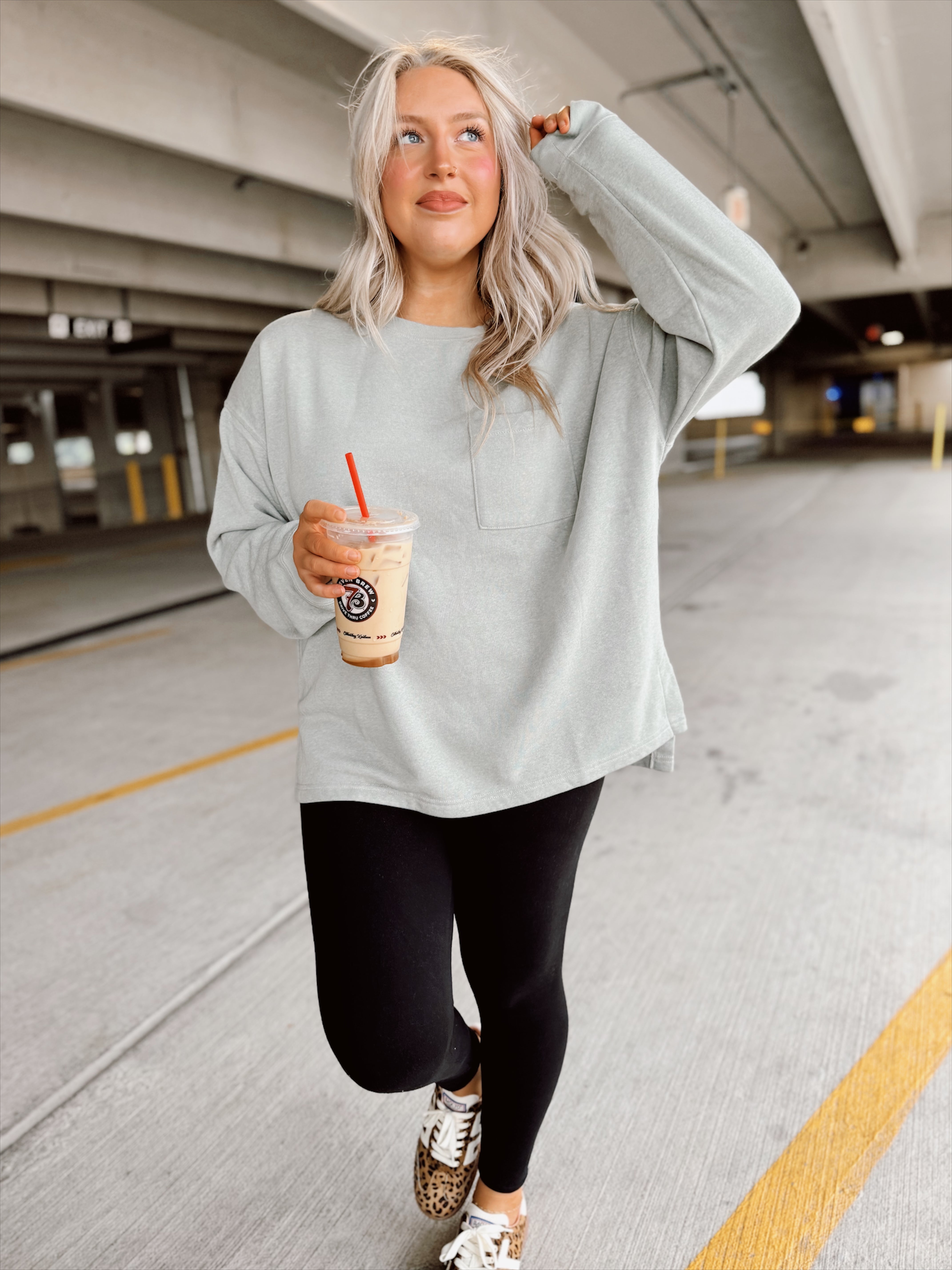 Woman in light sage sweatshirt and black leggings holding a drink in a parking garage.