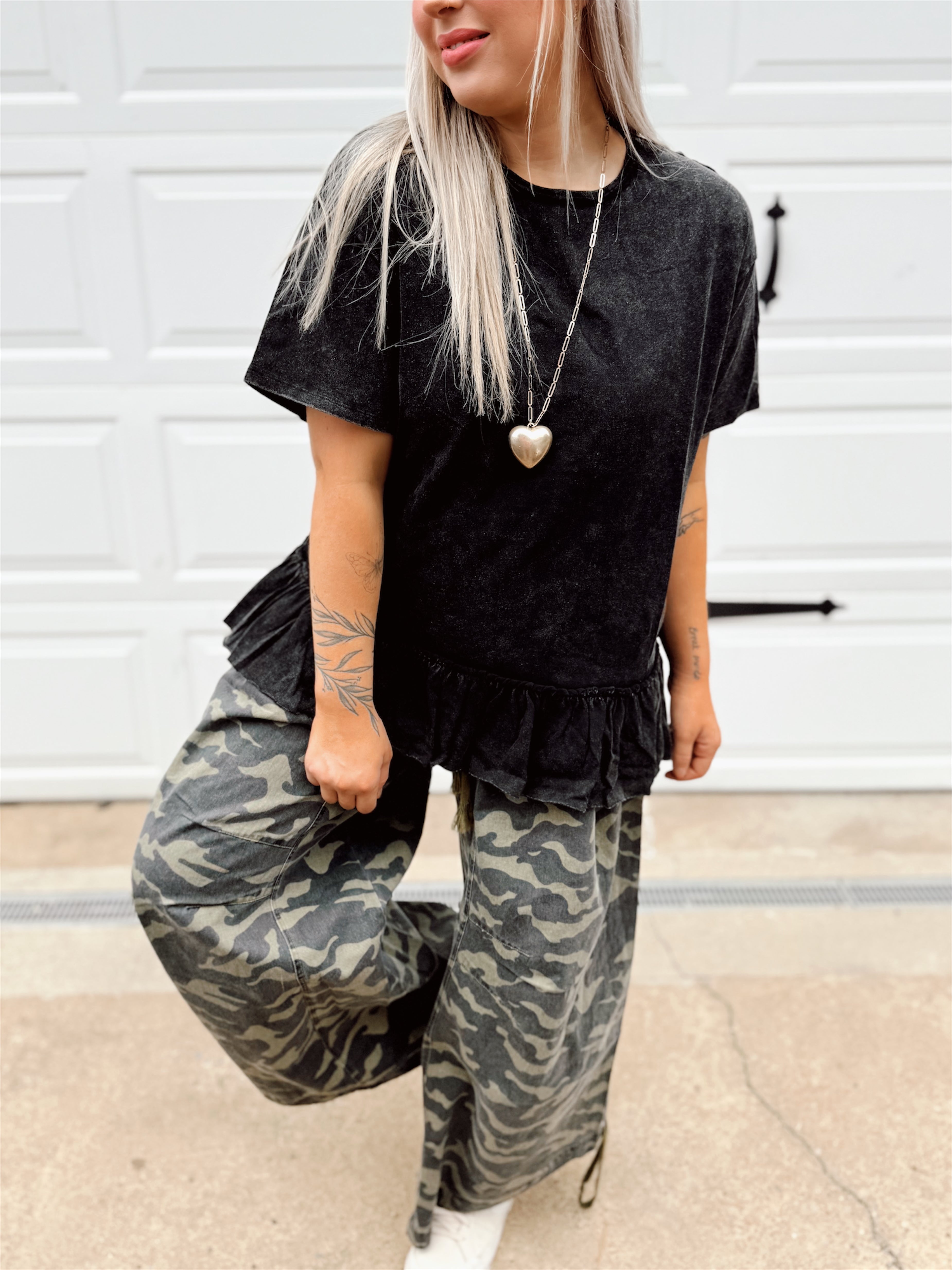 Person wearing a black ruffled peplum top and camouflage pants standing in front of a white wall.