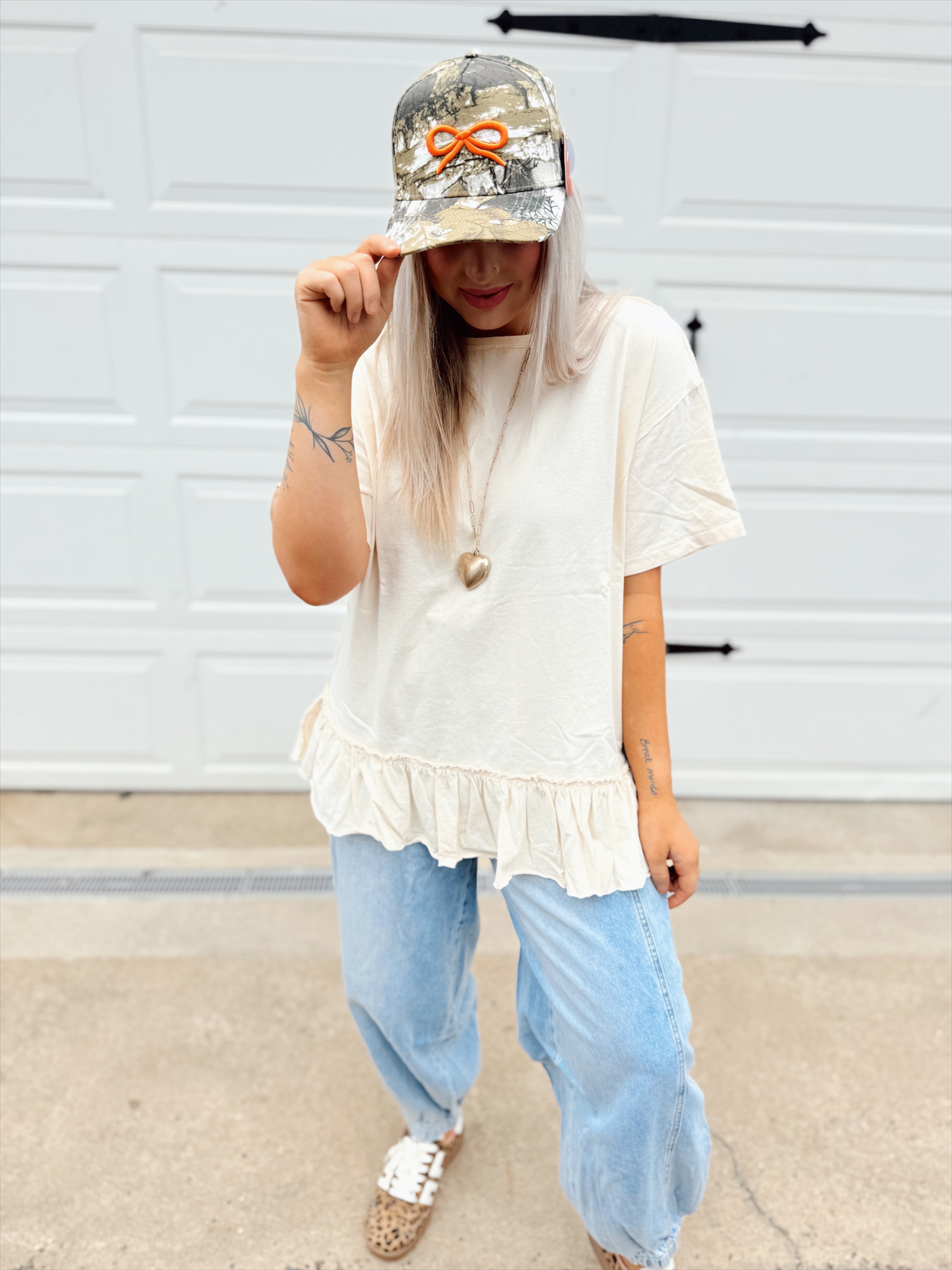 Person wearing a white top with ruffled hem, light blue jeans, and a camouflage cap with an orange design.