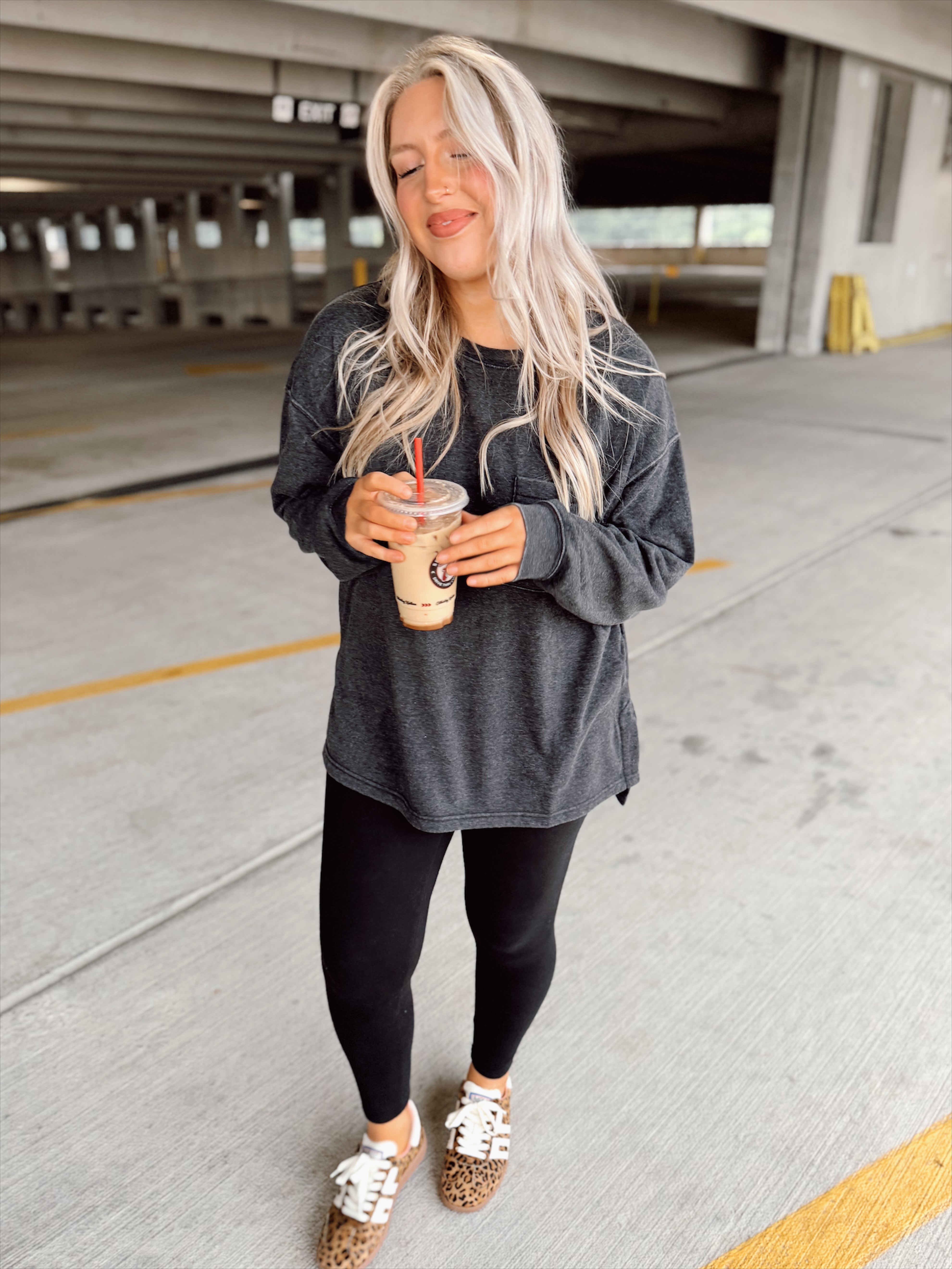 Woman in black sweatshirt and black leggings holding a drink in an parking garage.