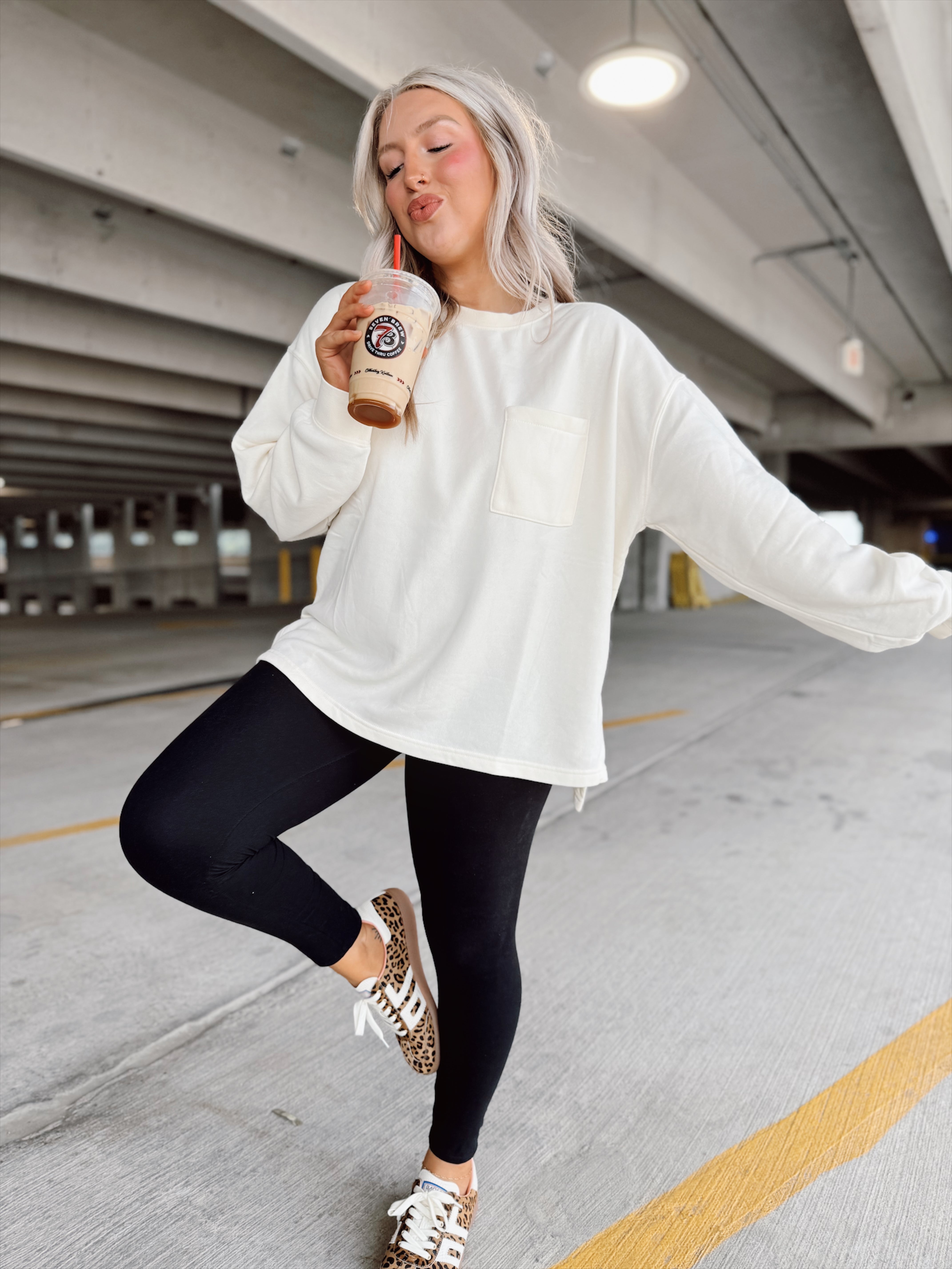 Person in a white sweatshirt and black leggings posing in an indoor parking garage, holding a drink.