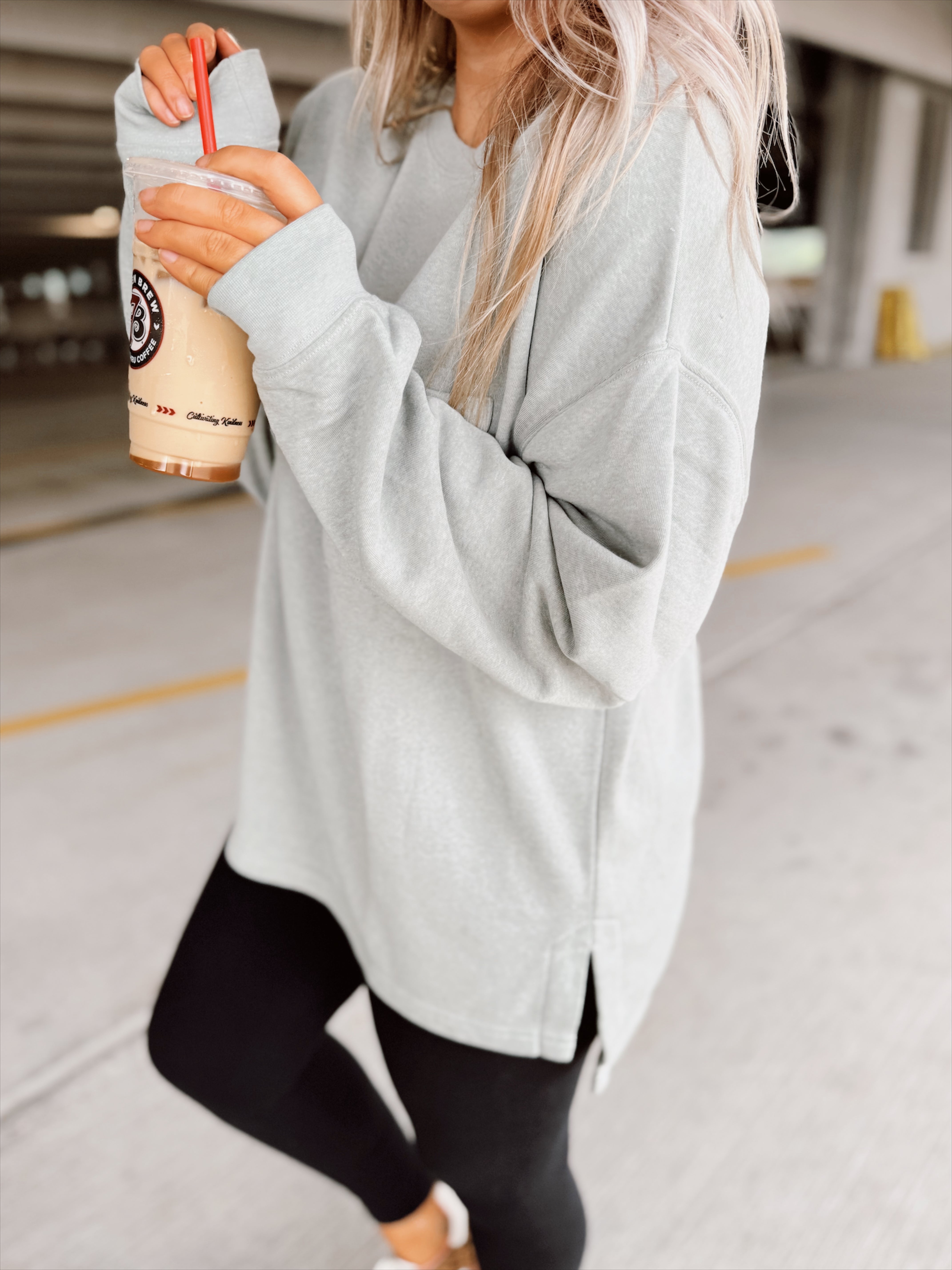 Woman in light sage sweatshirt and black leggings holding a drink in a parking garage.