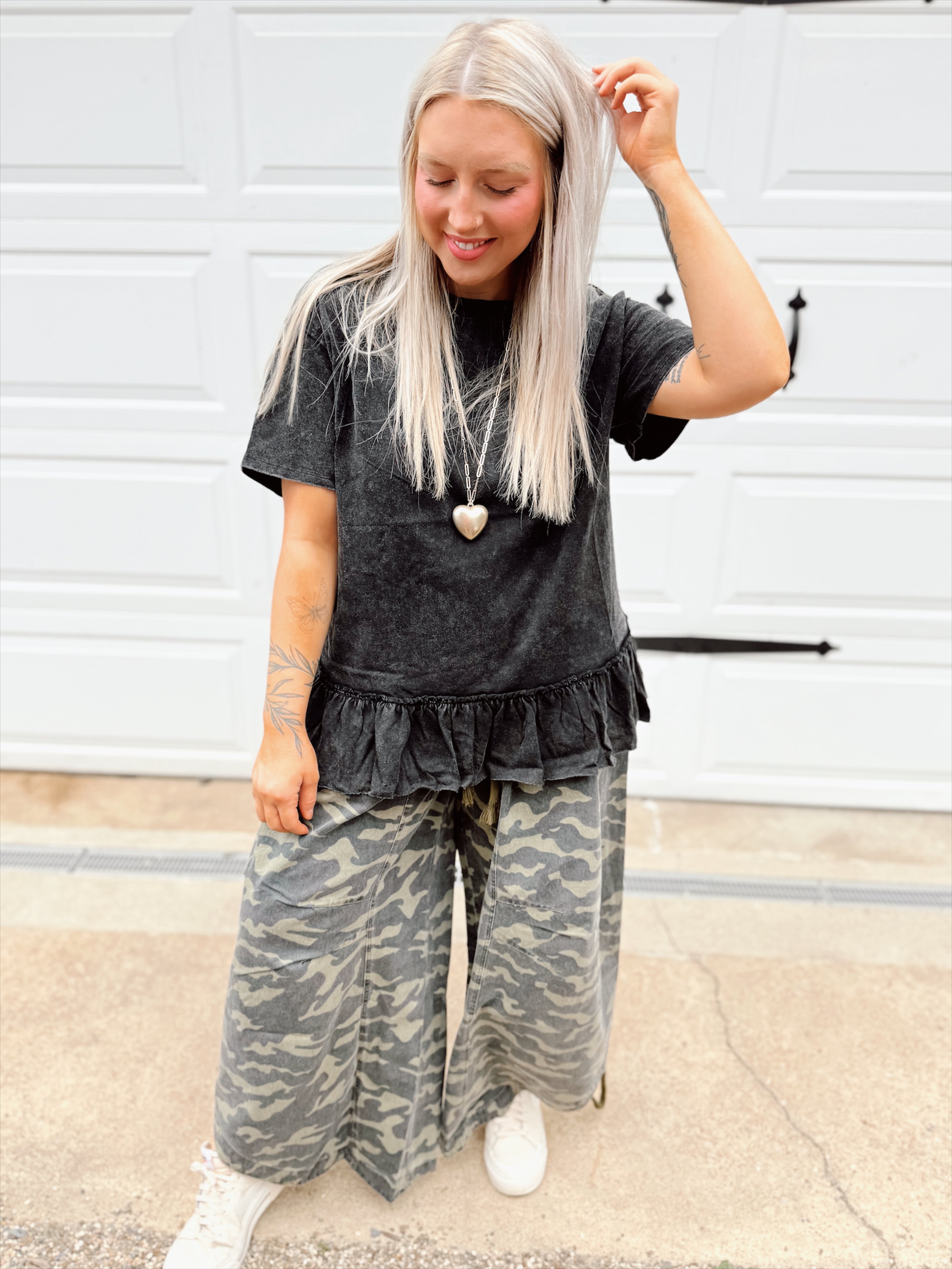 Person wearing a black ruffled peplum top and camouflage pants standing in front of a white wall.