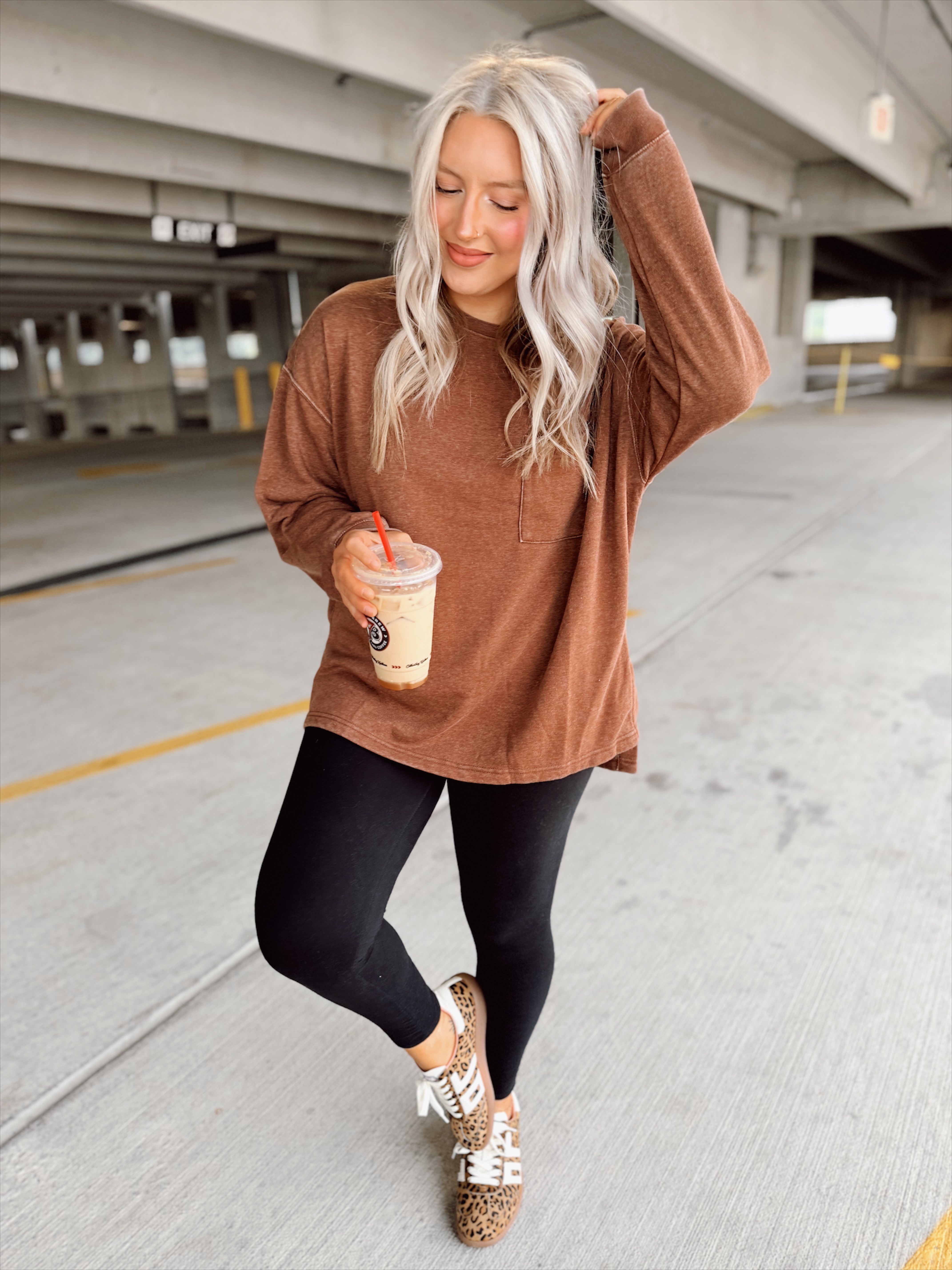 Woman in a brown sweater and black leggings holding a drink in an indoor parking garage.