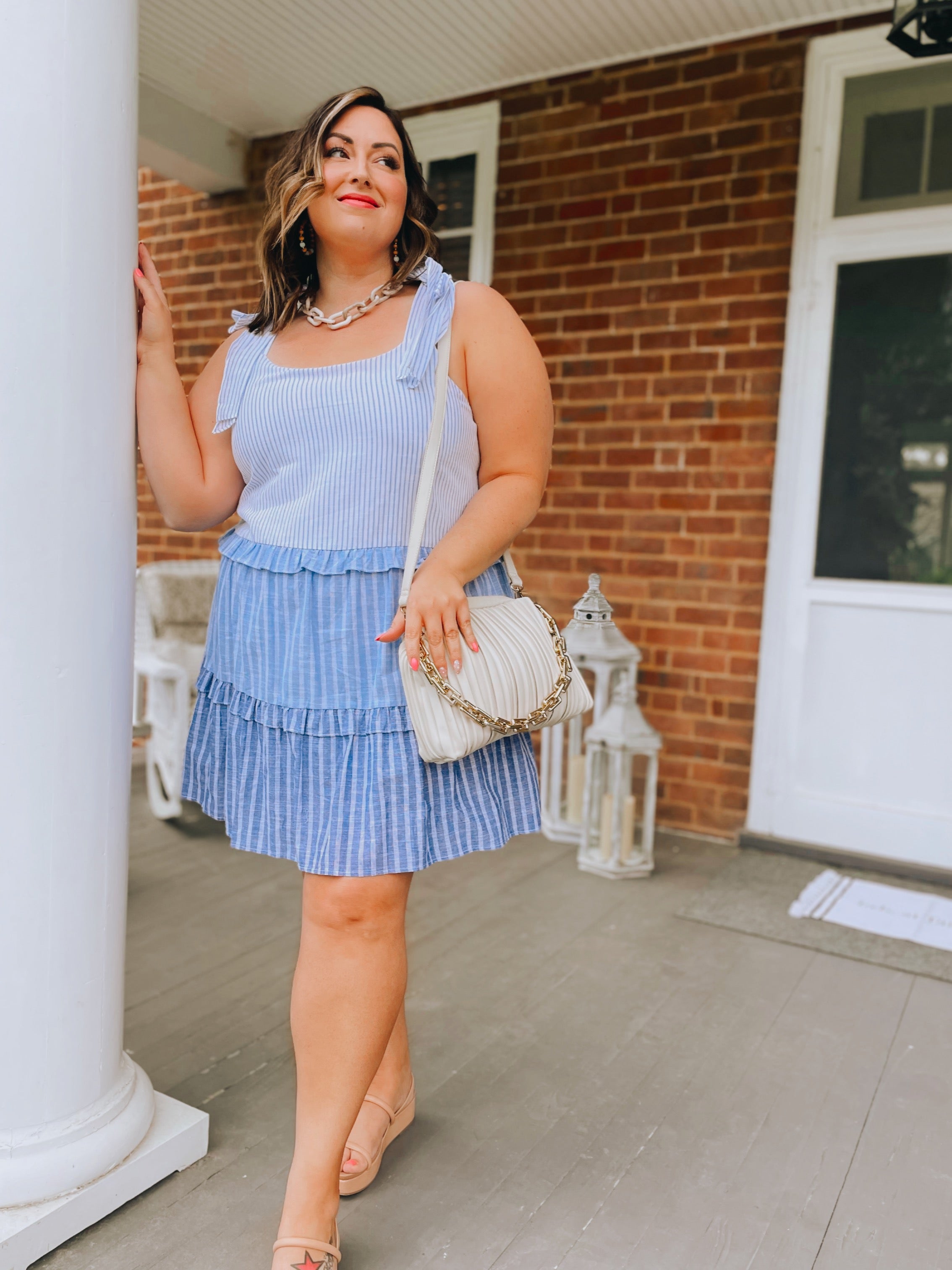 A woman in a blue and white striped dress, nude sandals, and the Rylee Clutch in oatmilk with a chunky gold chain stands on a brick porch, smiling and leaning against a white column. Decorative lanterns are placed behind her.