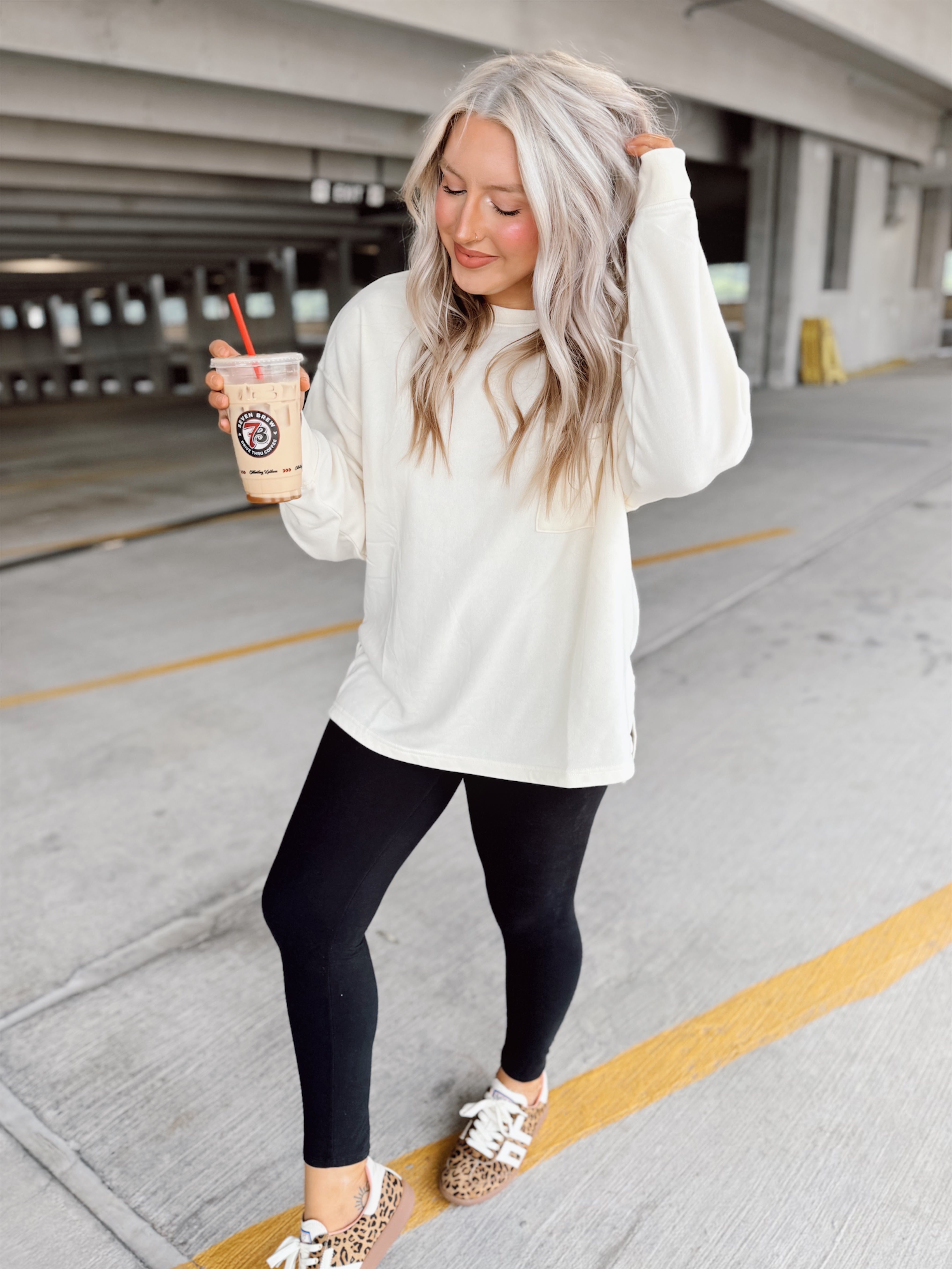 Woman in a white sweatshirt and black leggings holding a drink in a parking garage.