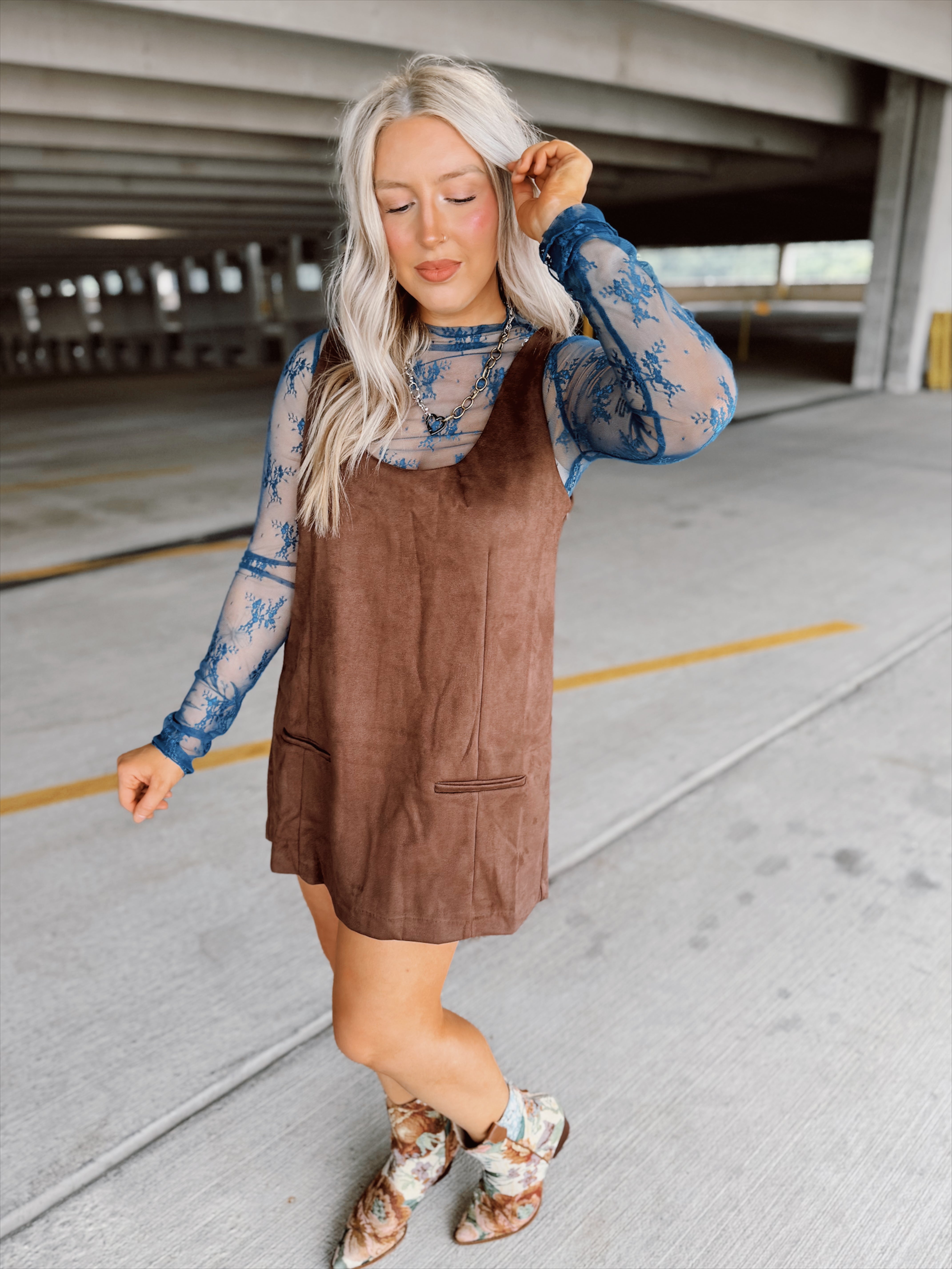 Woman in a brown suede dress with floral top and boots standing in a parking garage.