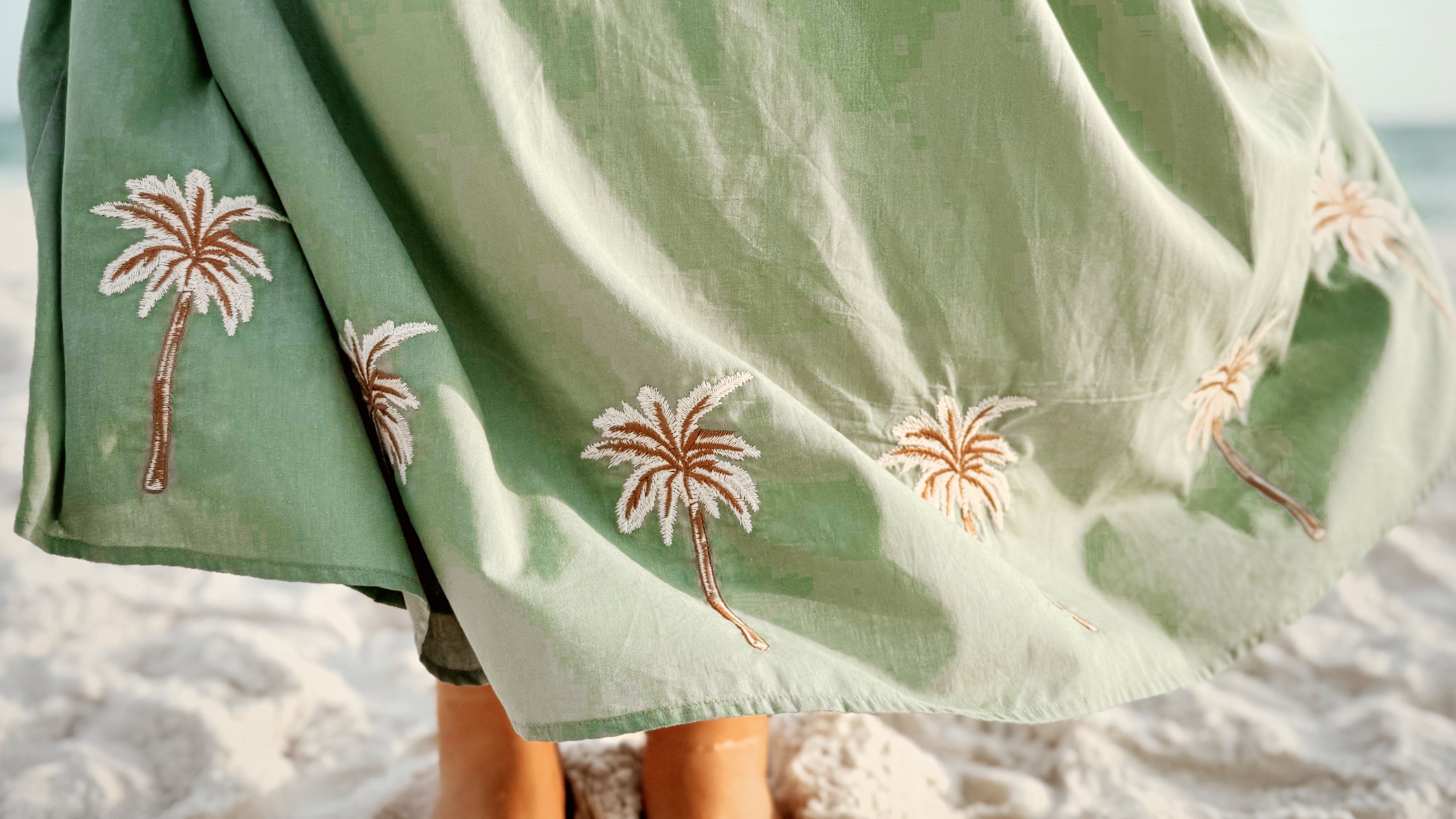 Person wearing a green dress with palm tree embroidery on a beach.