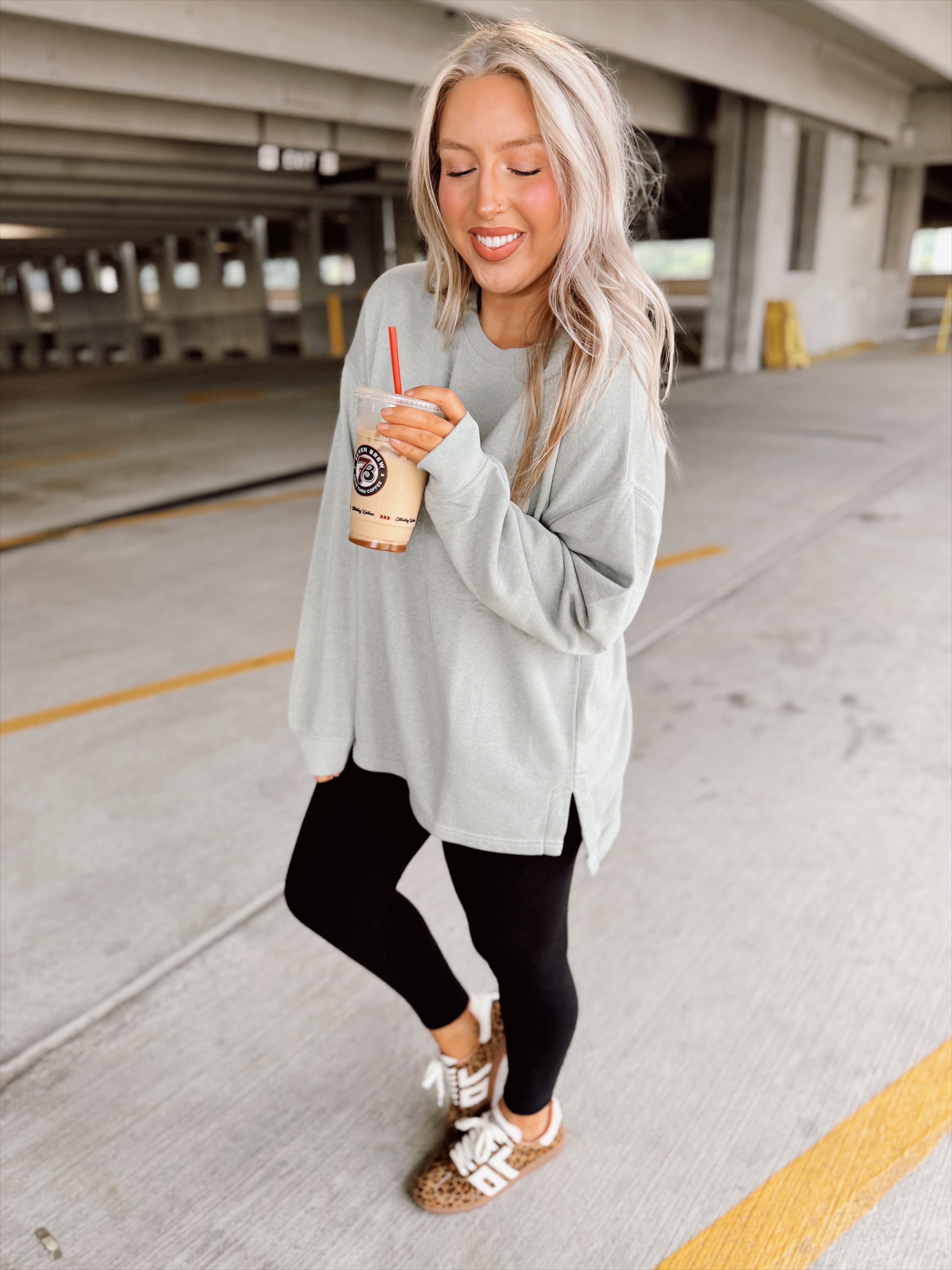 Woman in light sage sweatshirt and black leggings holding a drink in a parking garage.