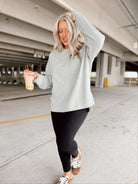 Woman in light sage sweatshirt and black leggings holding a drink in a parking garage.