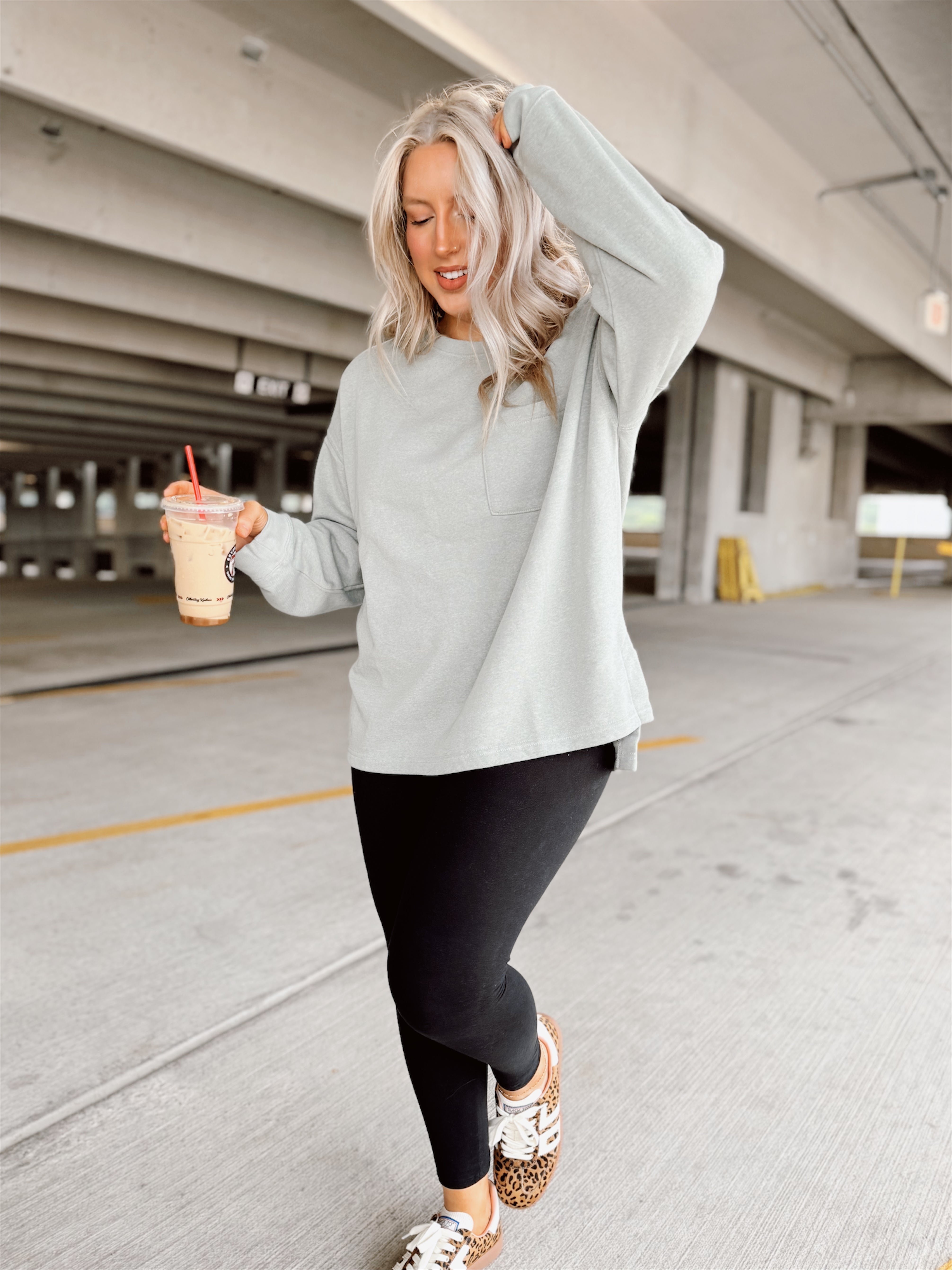 Woman in light sage sweatshirt and black leggings holding a drink in a parking garage.