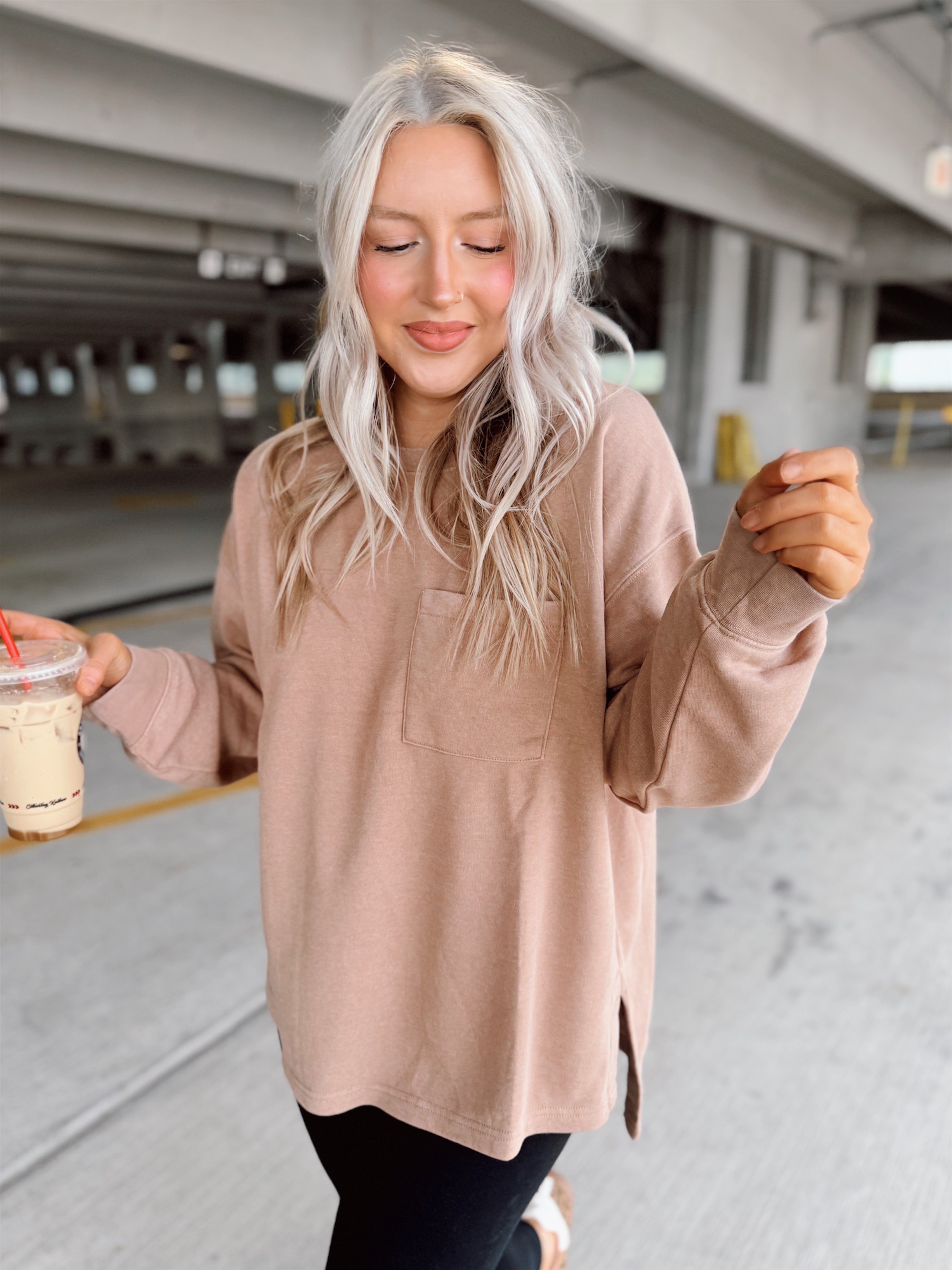 Woman in a beige sweater and black leggings holding a coffee cup in an underground parking lot.