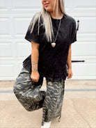 Person wearing a black ruffled peplum top and camouflage pants standing in front of a white wall.