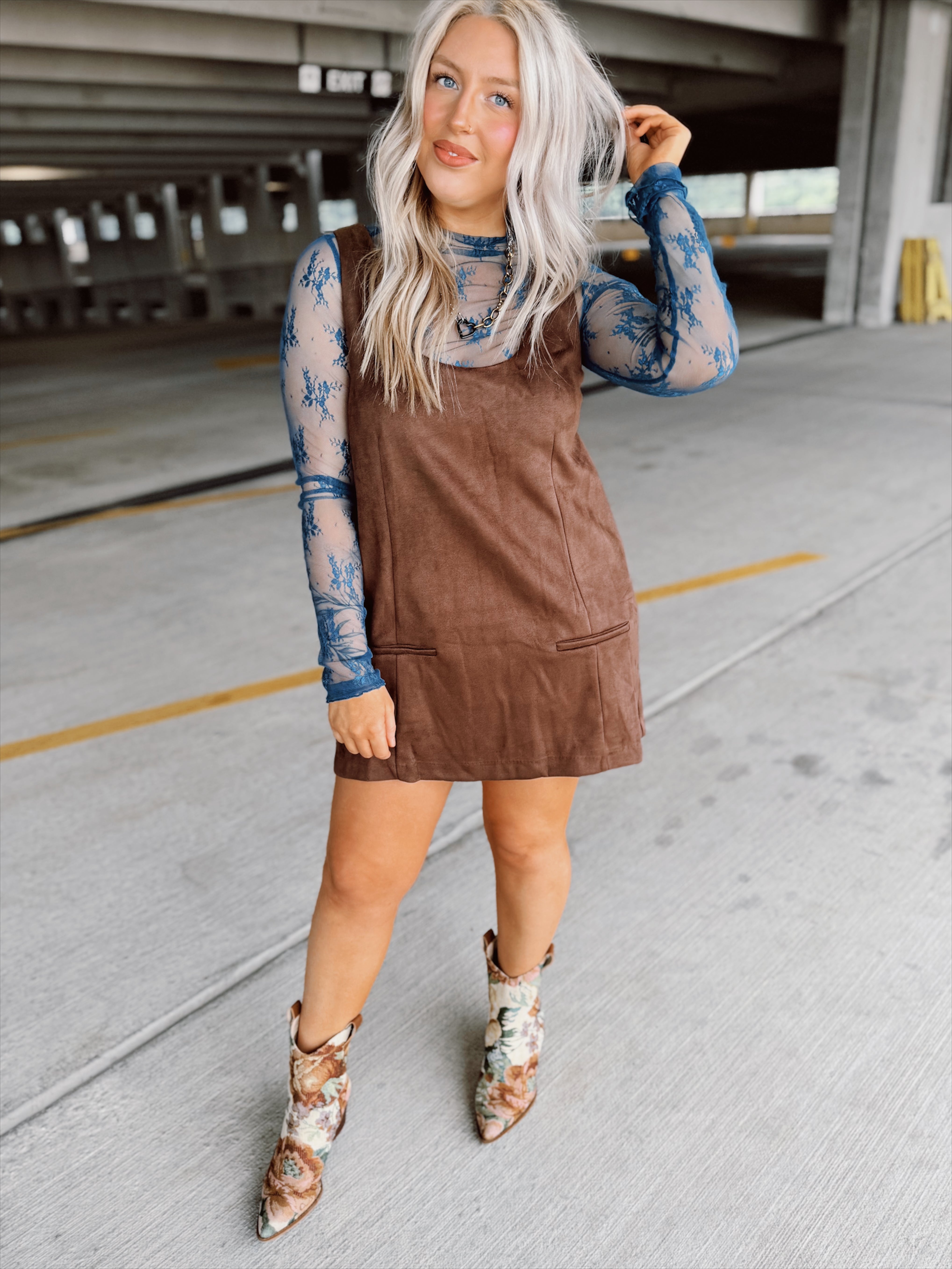Woman in a brown suede dress with floral top and boots standing in a parking garage.