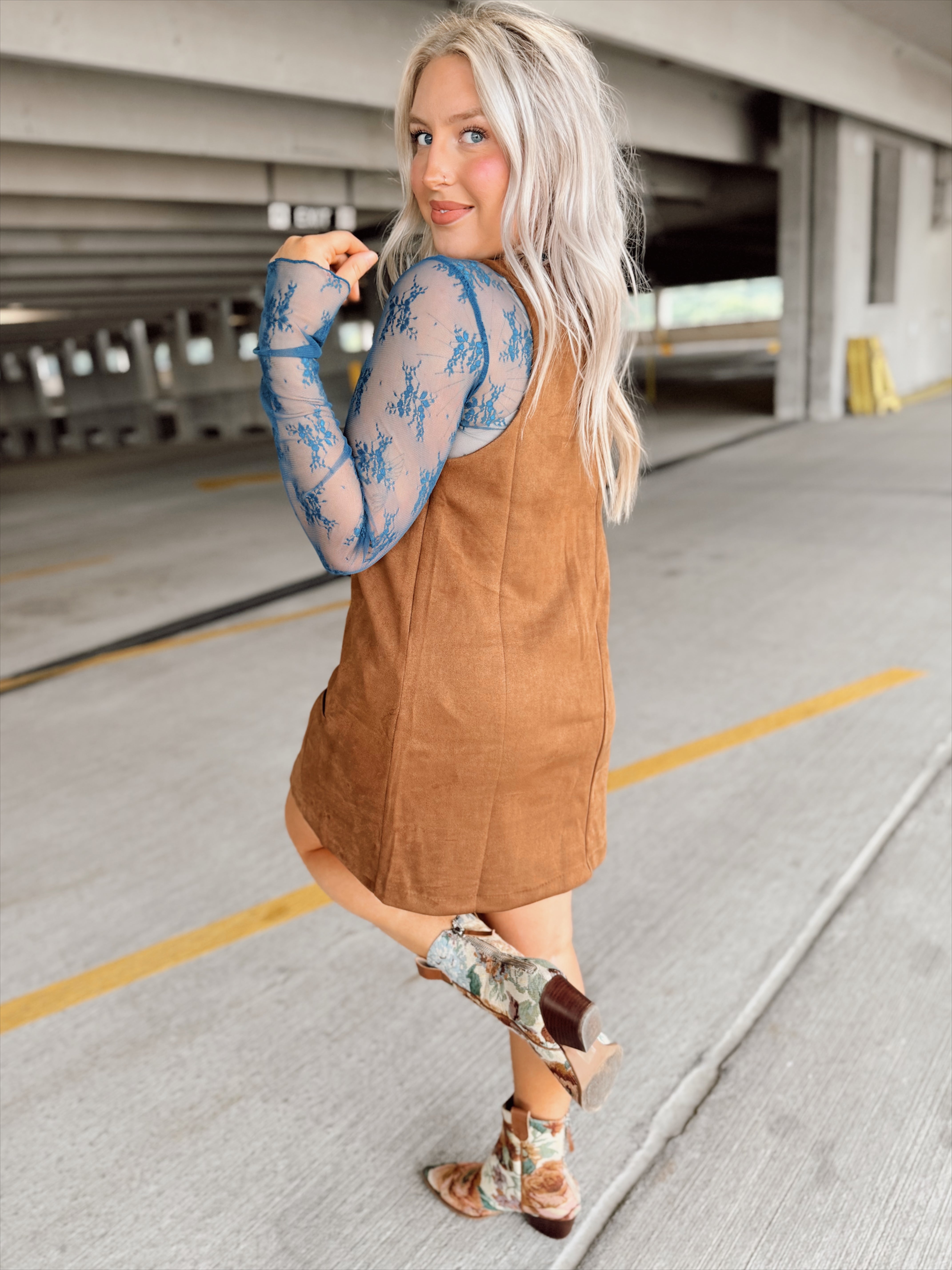 Woman in a brown suede dress with floral top and boots standing in a parking garage.