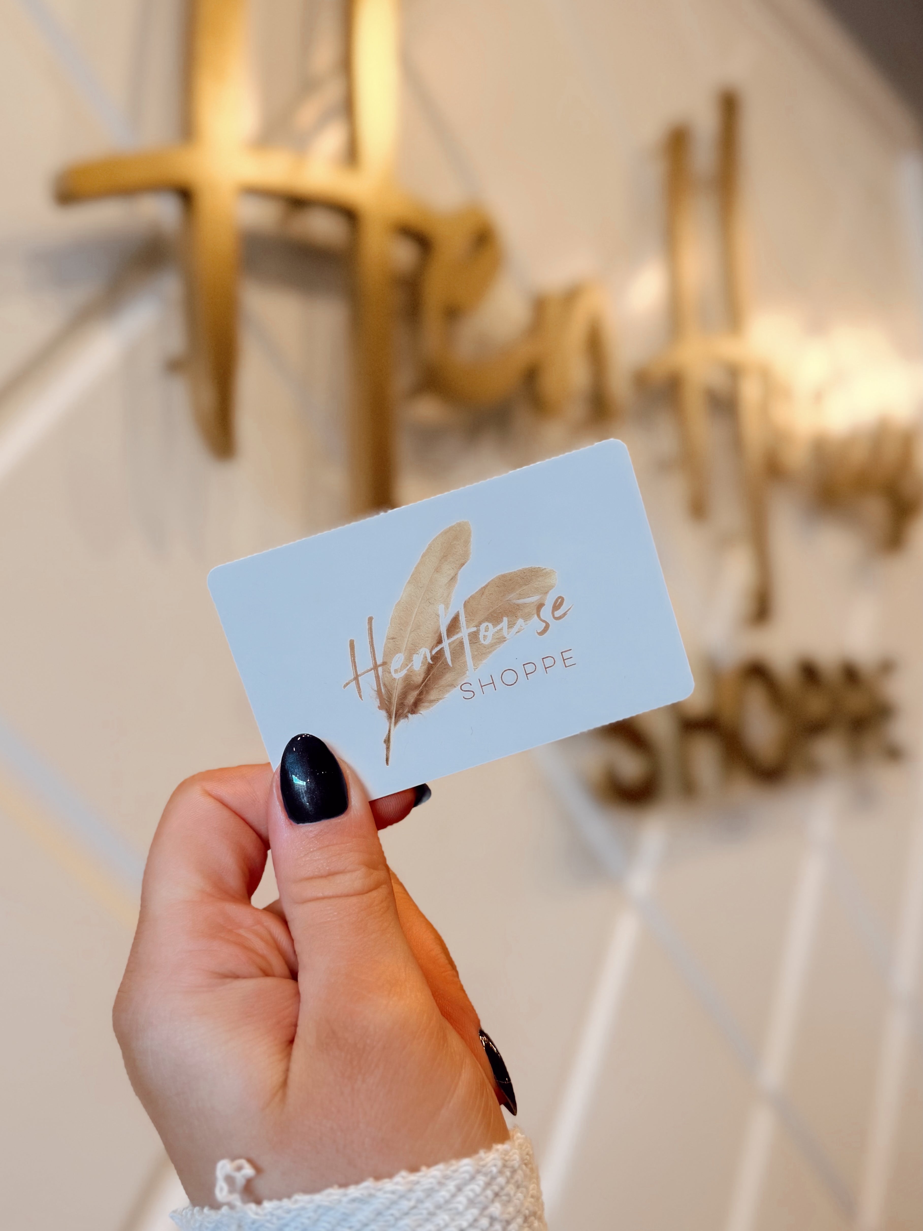 A hand with black nail polish holds a GIFT CARD in front of a blurred Hen House Shoppe sign on a patterned wall.