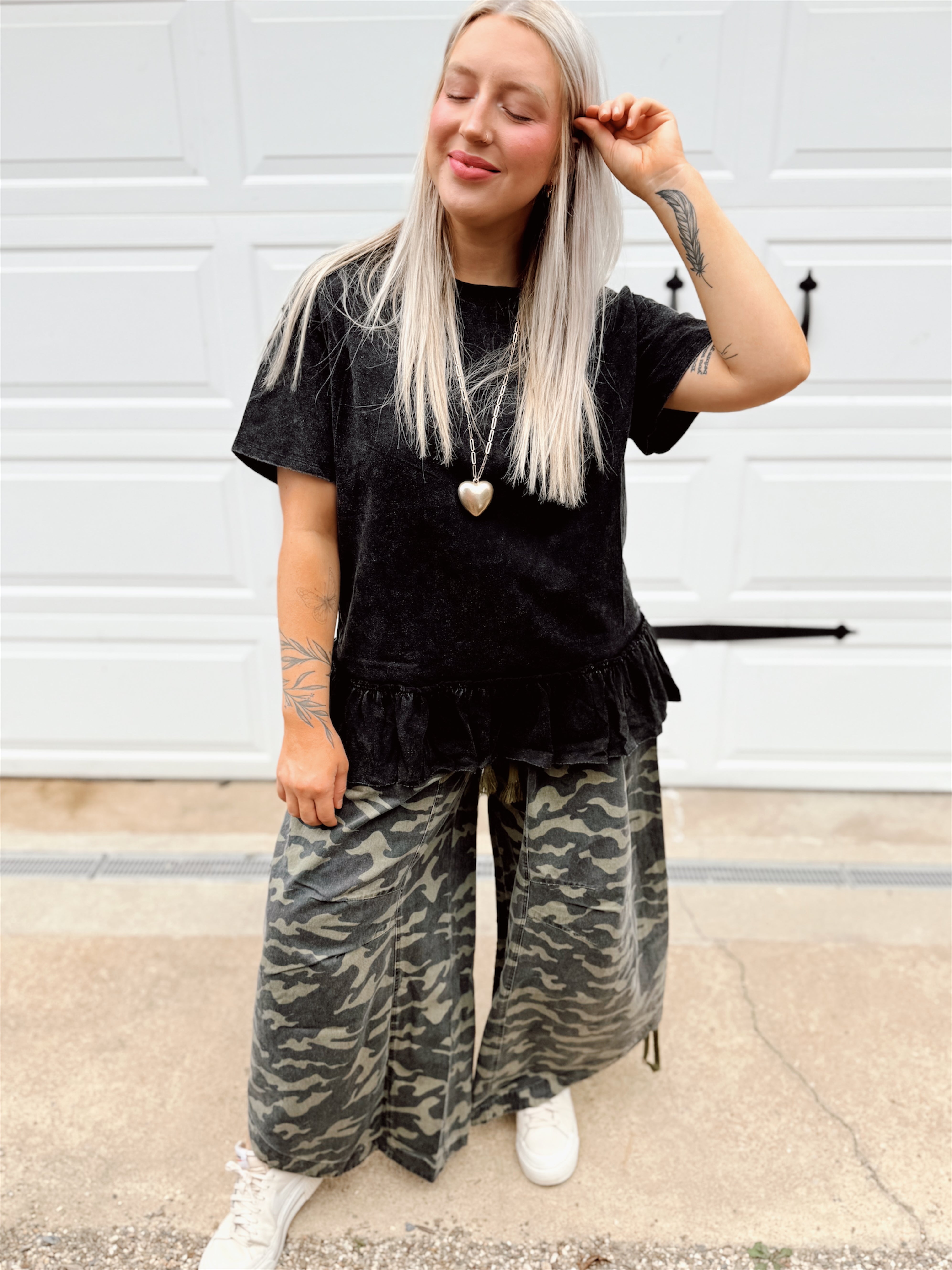 Person wearing a black ruffled peplum top and camouflage pants standing in front of a white wall.