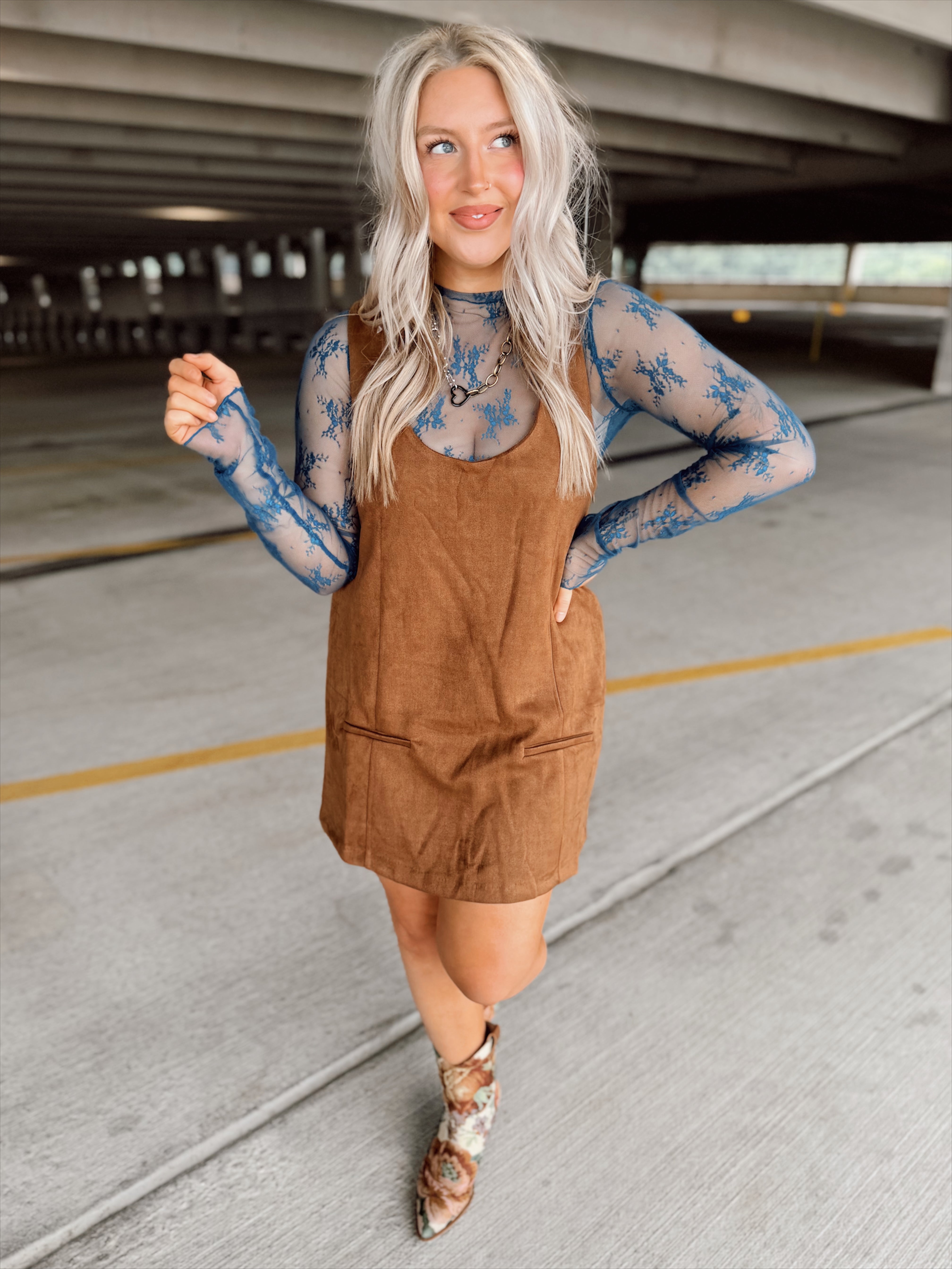 Woman in a brown suede dress with floral top and boots standing in an parking garage.
