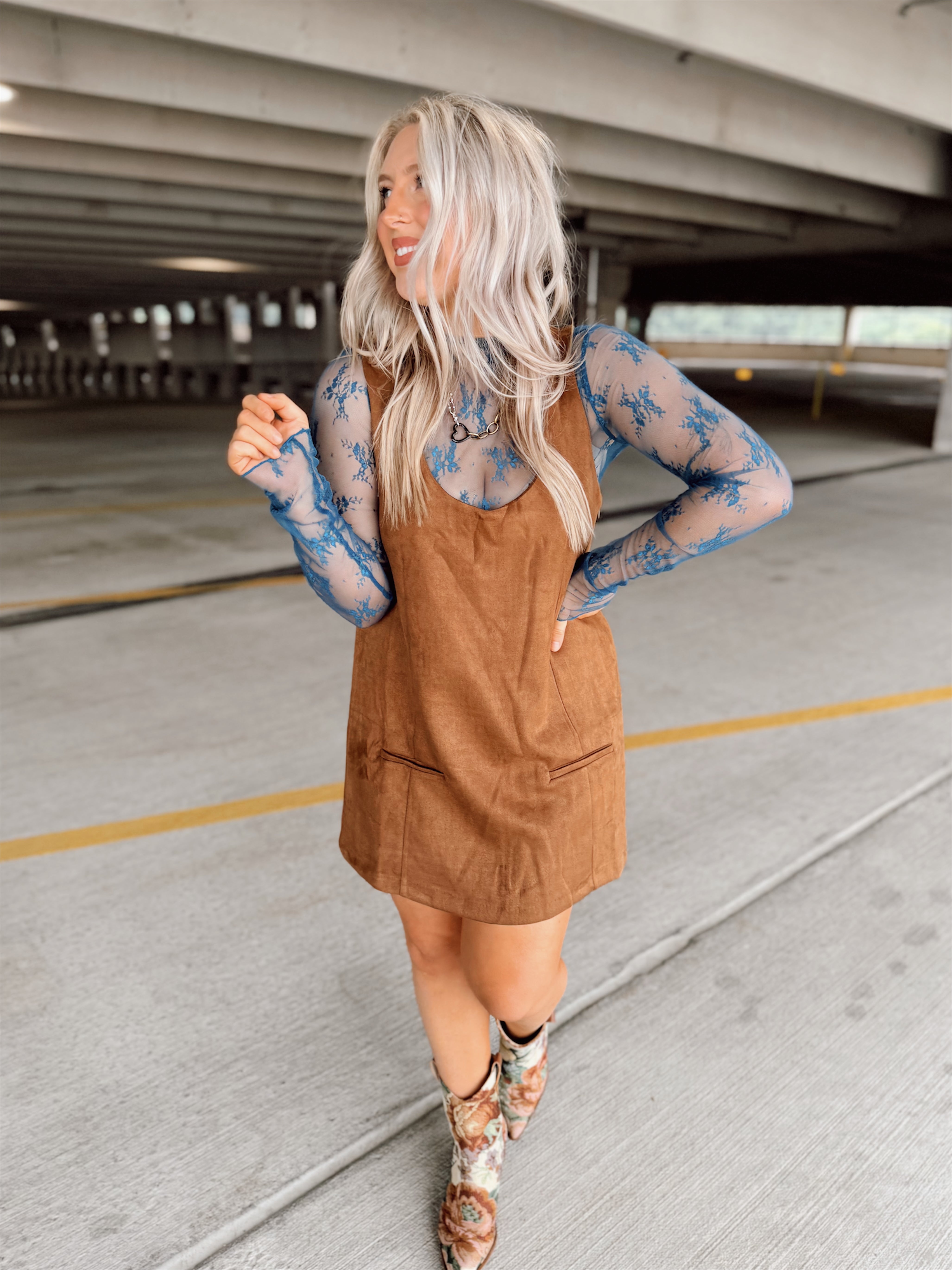 Woman in a brown suede dress with floral top and boots standing in an parking garage.