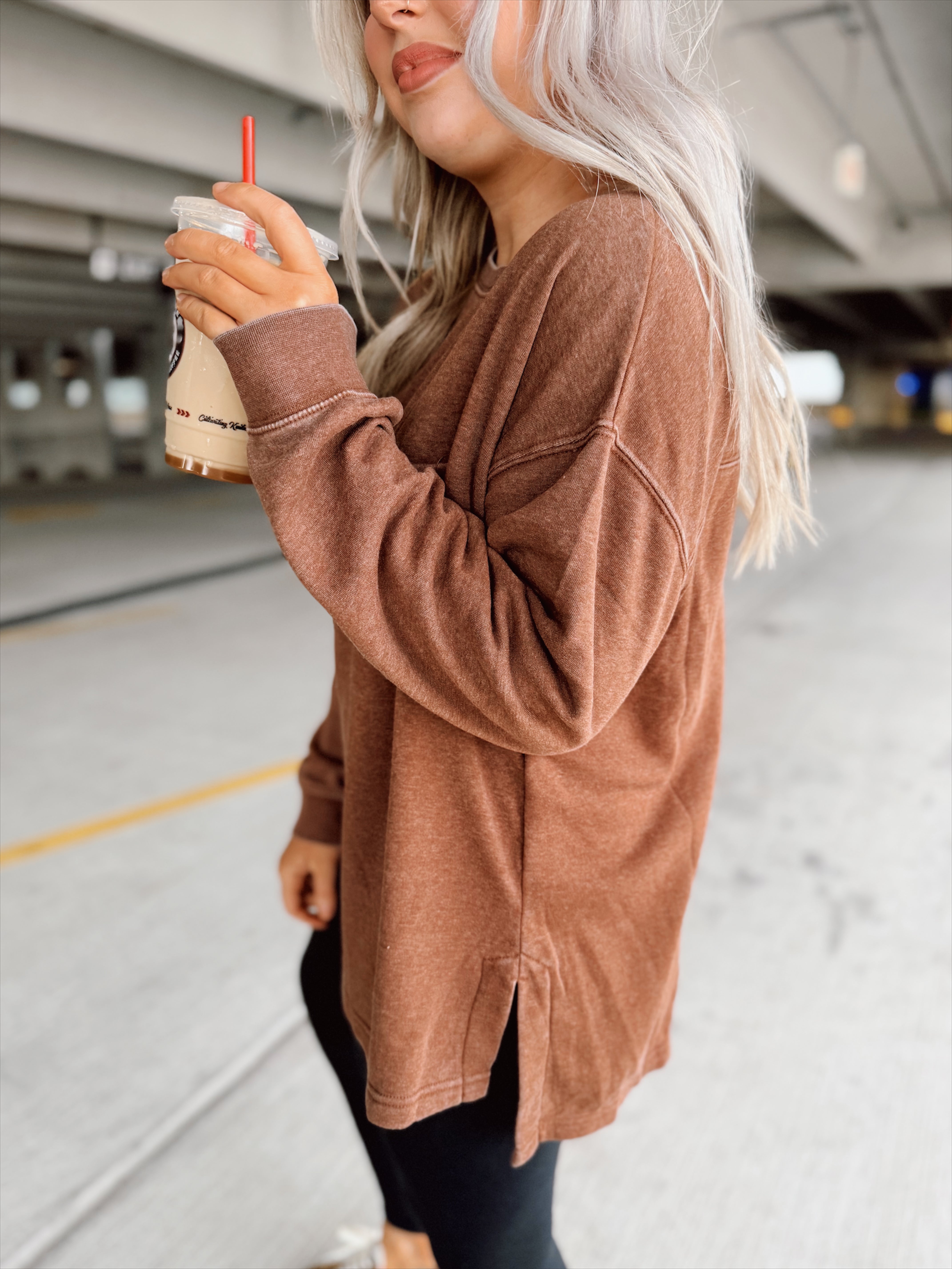 Woman in a brown oversized sweatshirt holding a drink in a parking garage.