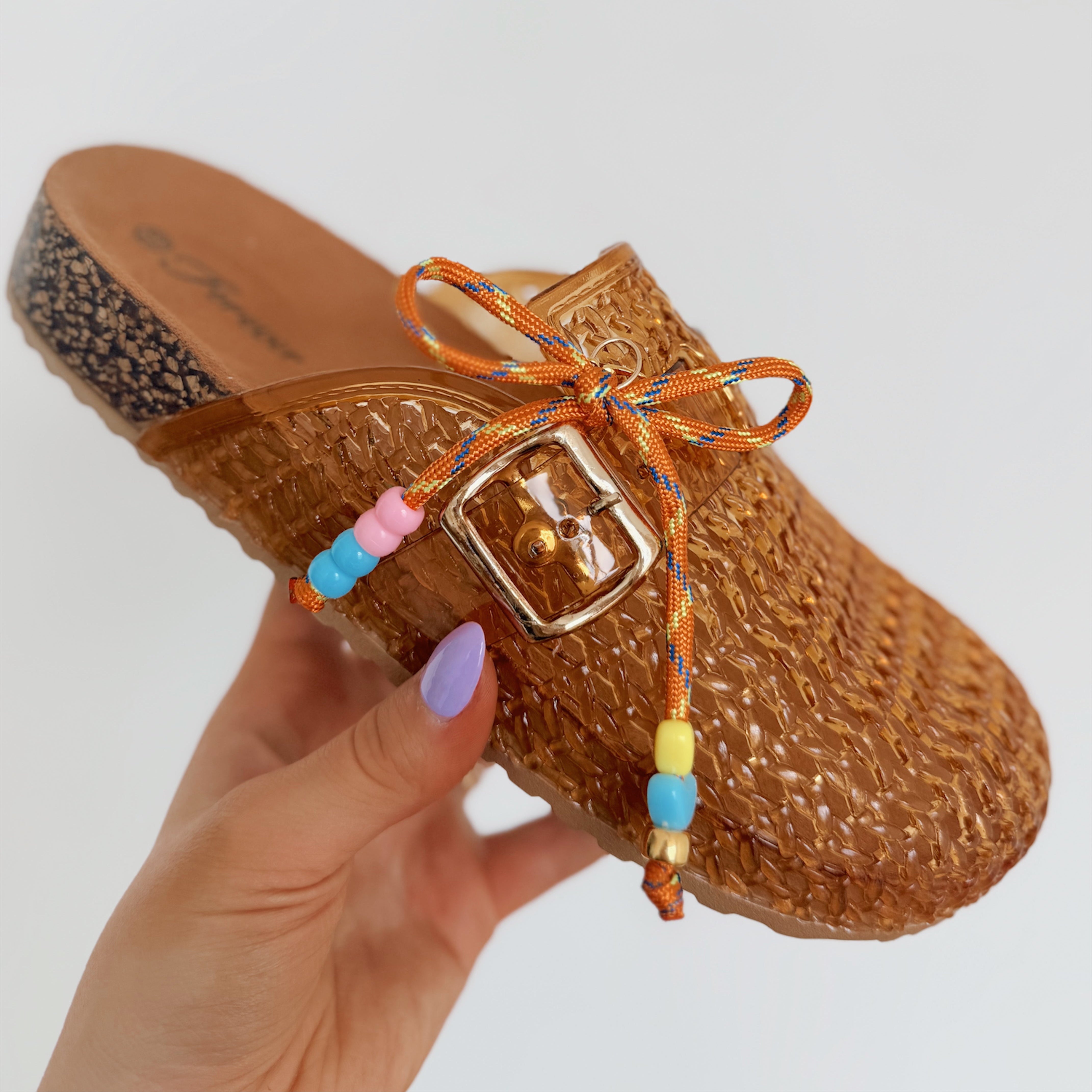Brown woven shoe with colorful beaded strap held by a hand on a light gray background