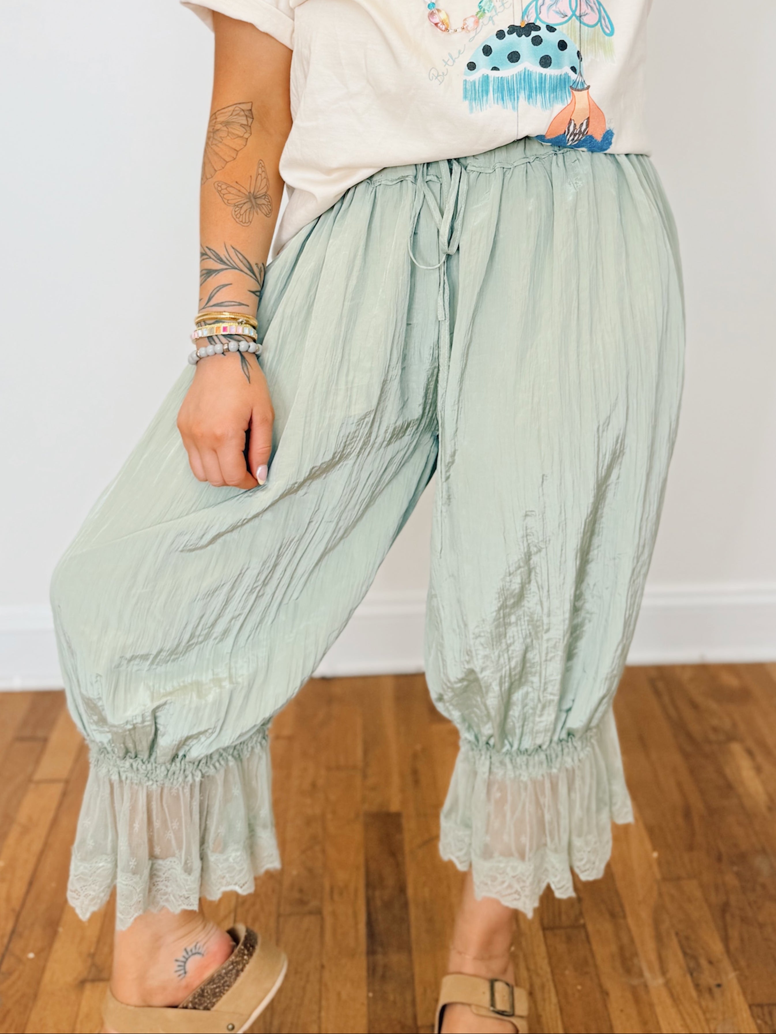 Person wearing a white t-shirt with colorful graphics and light green ruffled pants on a wooden floor.