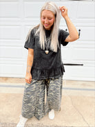 Person wearing a black ruffled peplum top and camouflage pants standing in front of a white wall.