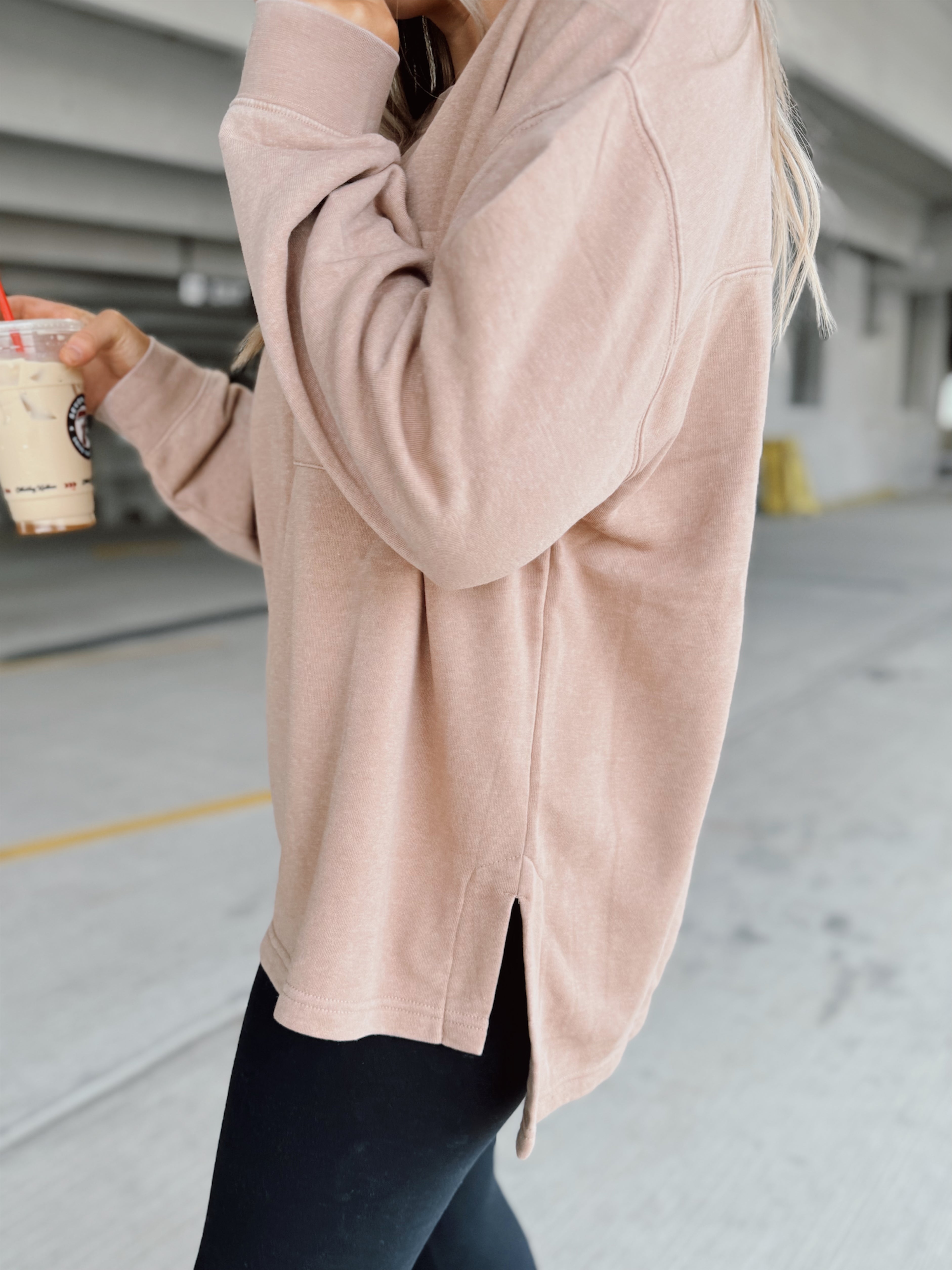 Woman in a beige sweater and black leggings holding a coffee cup in an underground parking lot.