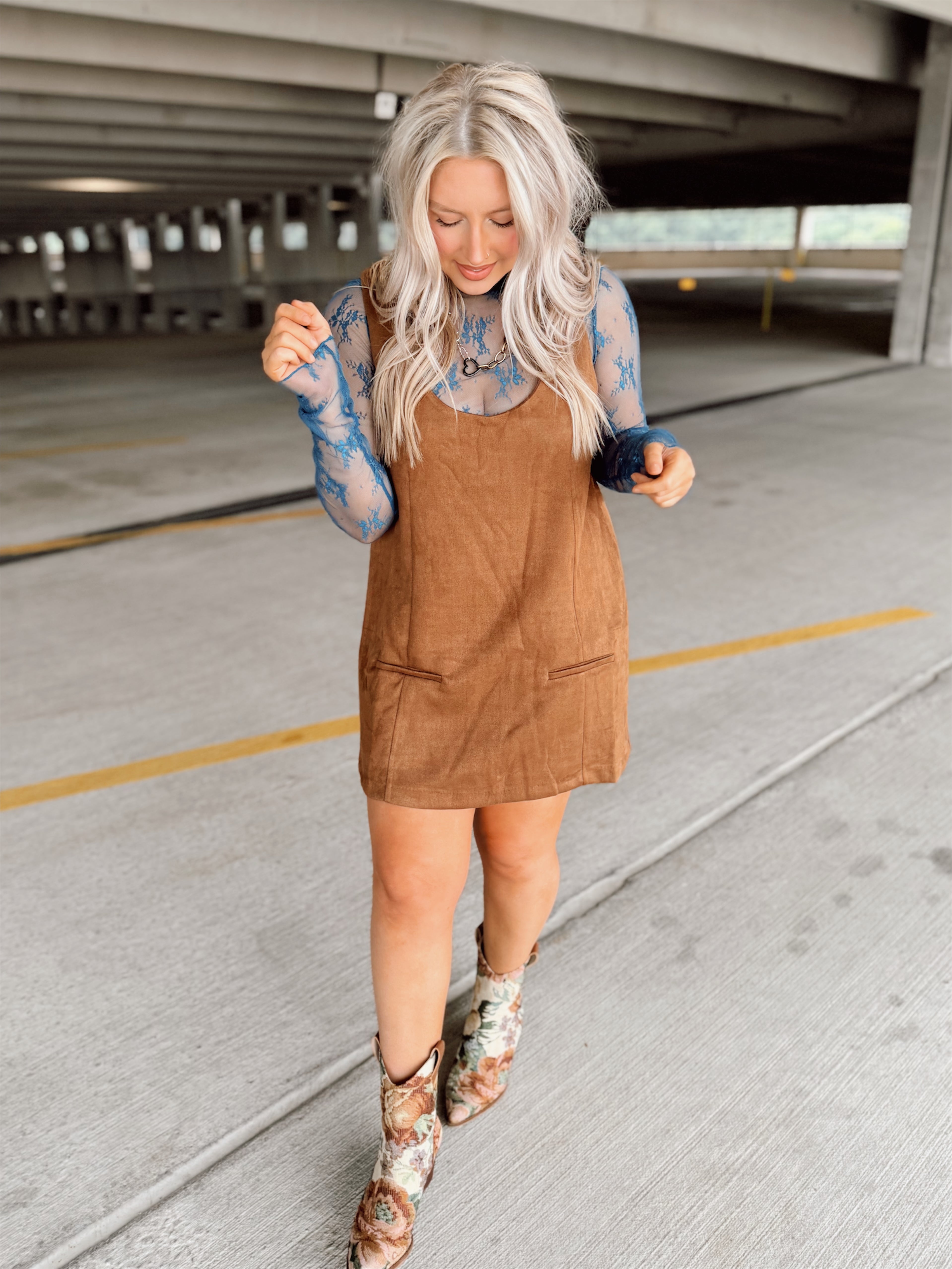 Woman in a brown suede dress with floral top and boots standing in an parking garage.