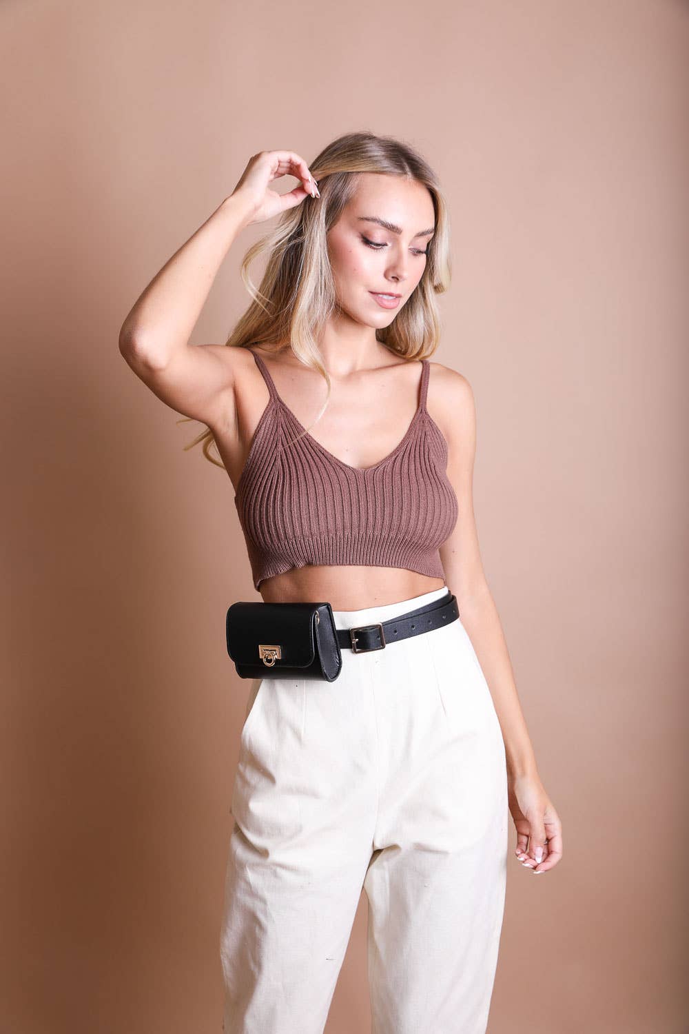 A woman with long blonde hair stands against a beige background, wearing a brown ribbed crop top, high-waisted cream pants, and the Mini Faux Leather Waist Belt Bag in black, with her left hand raised near her head.