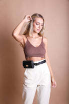 A woman with long blonde hair stands against a beige background, wearing a brown ribbed crop top, high-waisted cream pants, and the Mini Faux Leather Waist Belt Bag in black, with her left hand raised near her head.