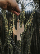 A hand holds up a black Desert Cactus Car Diffuser on a string, with green leafy branches and a wooden fence blurred in the background.