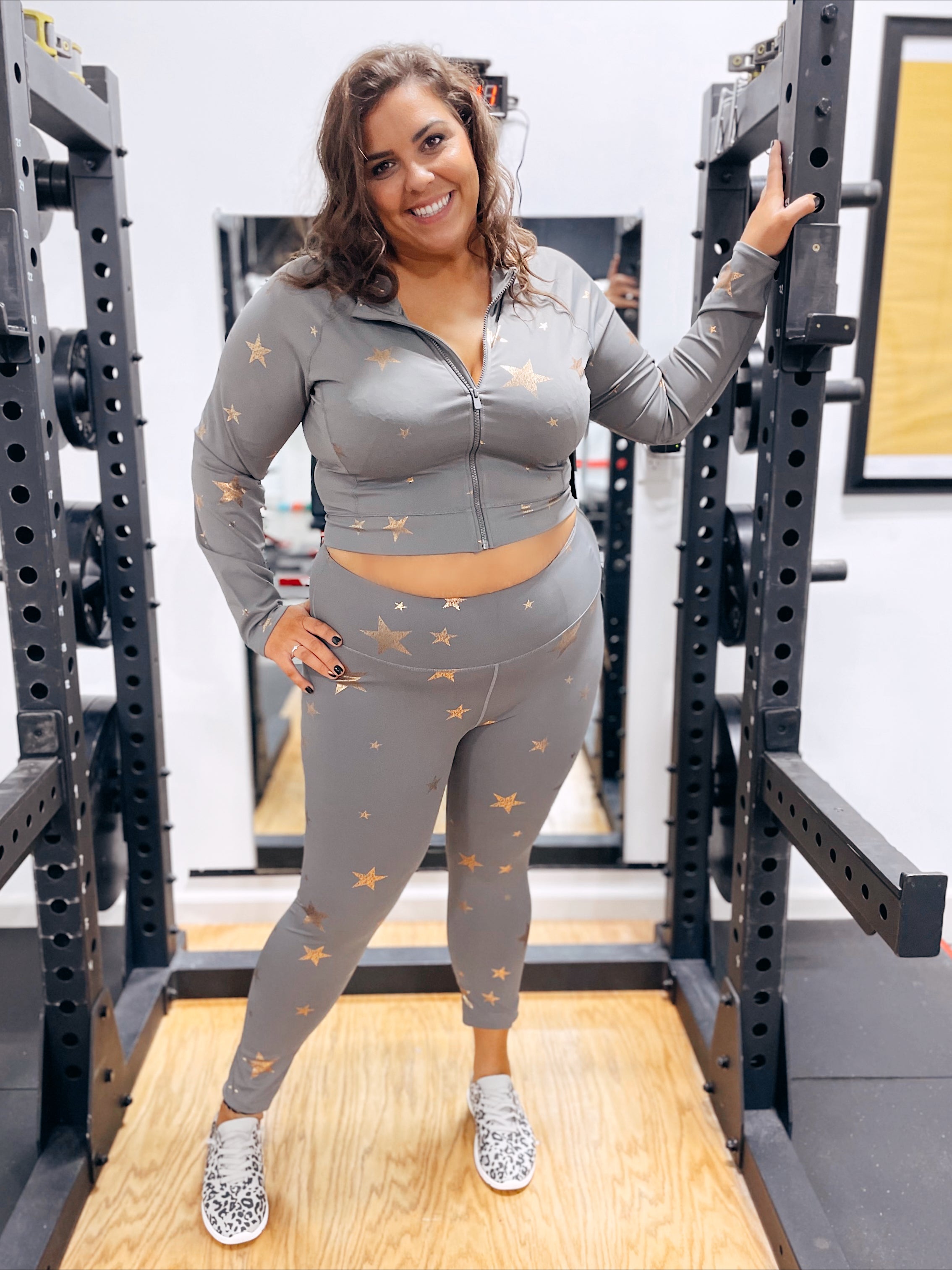 A woman with wavy brown hair smiles in a gym, holding a squat rack and wearing Curvy Starry Night Foiled Leggings—a gray, squat-proof athletic set with foil star print—paired with spotted slip-on sneakers.
