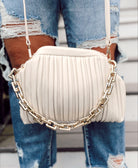 A person wearing ripped blue jeans holds the Rylee Clutch in oatmilk, featuring a pleated design, frame handle, chunky gold chain, and a detachable adjustable strap.