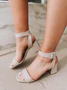 Close-up of a person wearing the Blest Smooth Heel in Beige by CL by Laundry—light beige, open-toe block heel sandals with ankle straps and buckles—standing on a concrete surface.