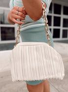 A person in a green and white striped dress holds the Rylee Clutch in oatmilk, featuring a chunky gold chain handle, against a backdrop of windows and a sidewalk.