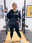 A woman wears Criss Cross Butter Leggings in Black and a zip-up workout top, holding a light blue kettlebell in her left hand as she stands confidently in a gym with exercise equipment and framed photos on the walls.