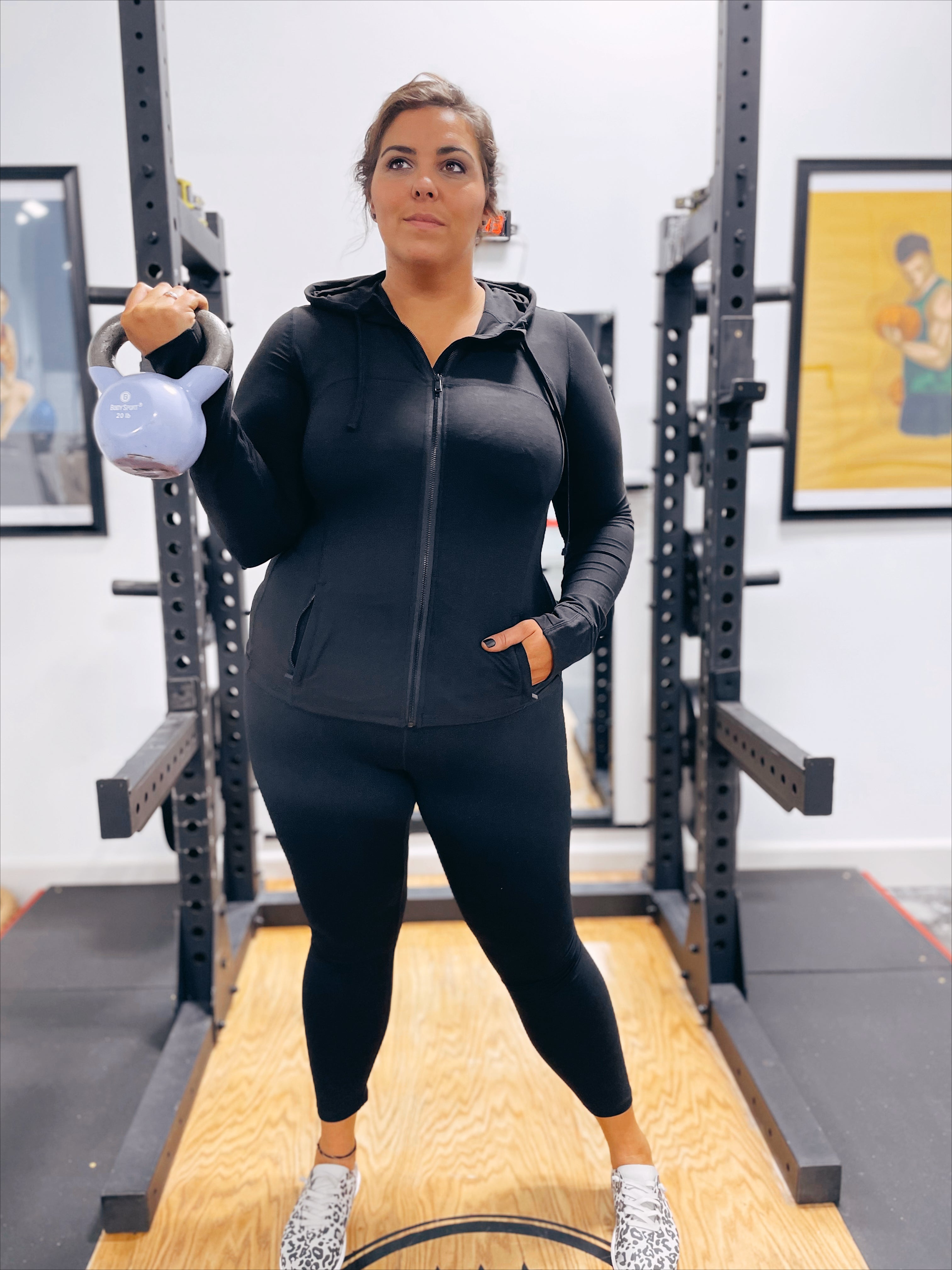A woman wears Criss Cross Butter Leggings in Black and a zip-up workout top, holding a light blue kettlebell in her left hand as she stands confidently in a gym with exercise equipment and framed photos on the walls.