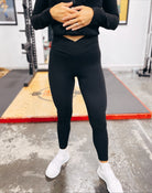 At the gym, a person wears the Criss Cross Butter Leggings in Black with a matching long-sleeve top and white sneakers, hands resting on their midsection. Gym equipment can be seen in the background.