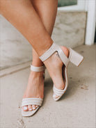 A close-up of a person wearing the Blest Smooth Heel in Beige by CL by Laundry—light beige, open-toe sandals with ankle straps and low block heels—standing on textured concrete with legs crossed.