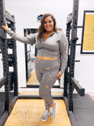 A woman smiles in a gym squat rack, wearing Curvy Starry Night Foiled Leggings with a gray star print athletic set and leopard print sneakers, confidently showing their squat-proof design.