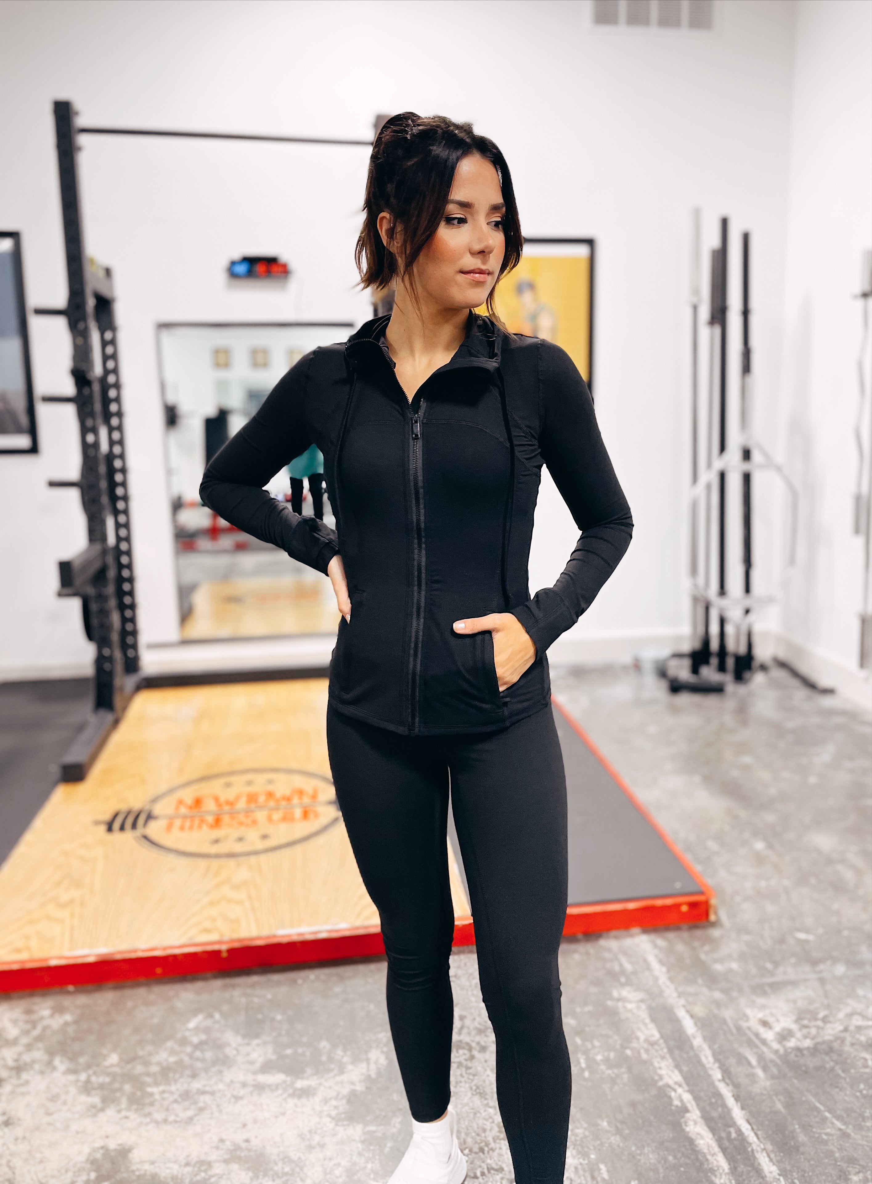 A woman stands confidently in a gym, hands in her jacket pockets, wearing Criss Cross Butter Leggings in Black. Gym equipment and a weightlifting platform appear in the background.