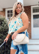 A woman with long, wavy blonde hair stands outdoors by steps and a railing, wearing a floral sleeveless top, ripped blue jeans, and carrying the Rylee Clutch in oatmilk. A brick building and greenery are in the background.