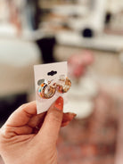 A hand with orange nail polish holds a white card featuring three pairs of Catriona earrings—small gold hoops with colorful enamel accents—against a blurred indoor background.