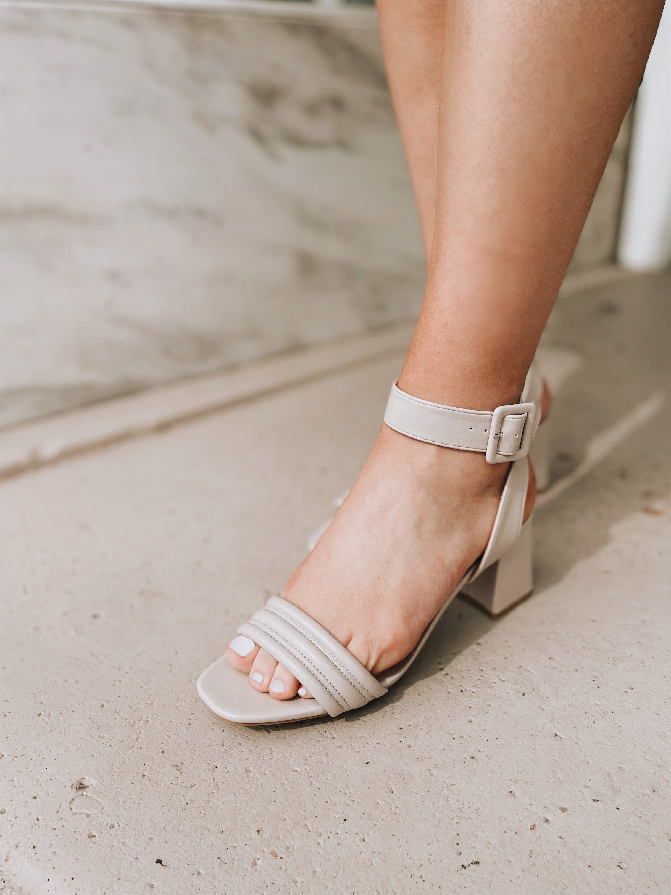 Someone wearing the Blest Smooth Heel in Beige by CL by Laundry—an open-toe block heel sandal with an ankle strap and buckle—stands on a light-colored stone or concrete surface.